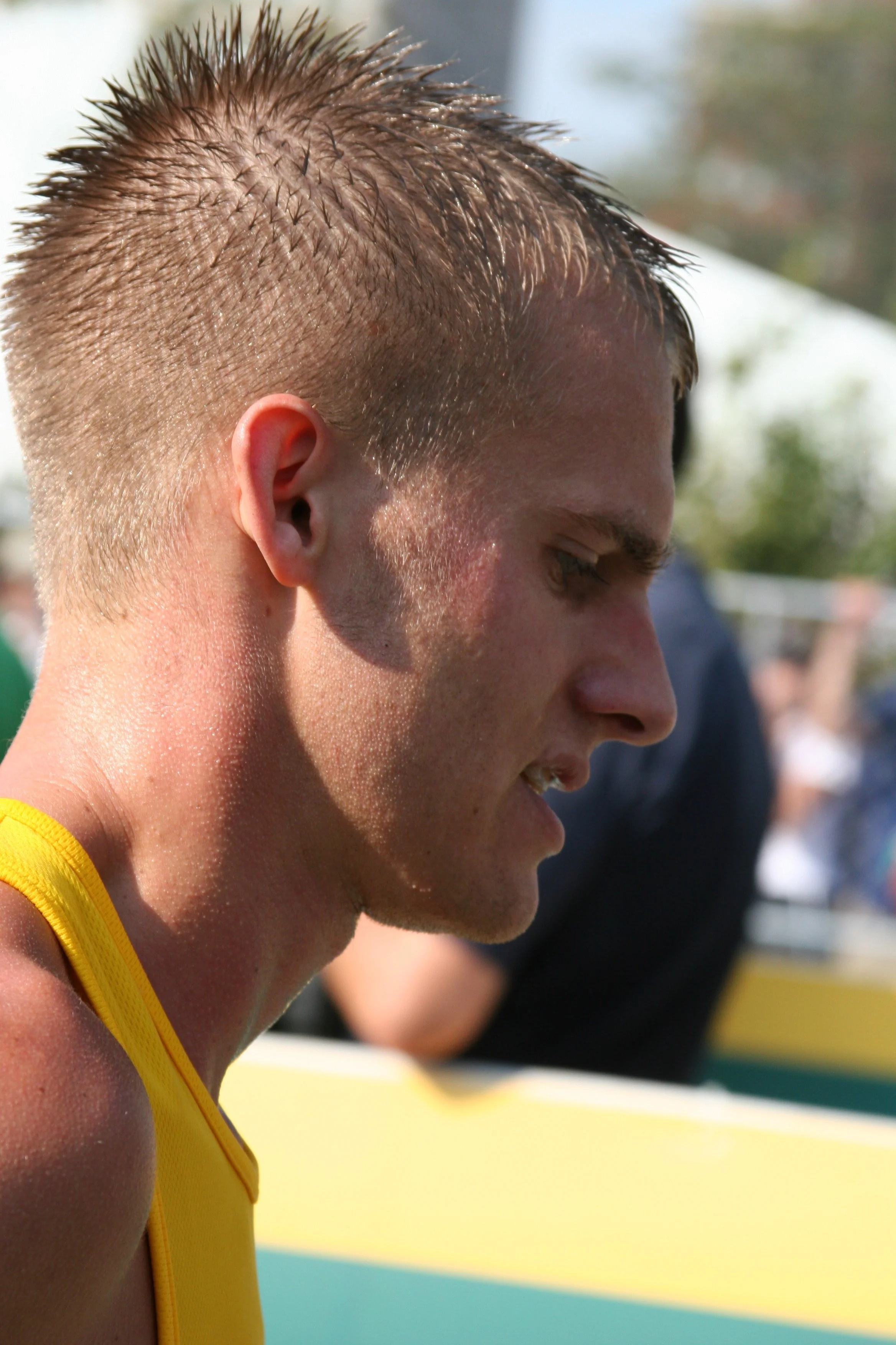 Close-up of a young male athlete with spiked blond hair wearing a yellow sports jersey, looking down, likely during a sports event outdoors.