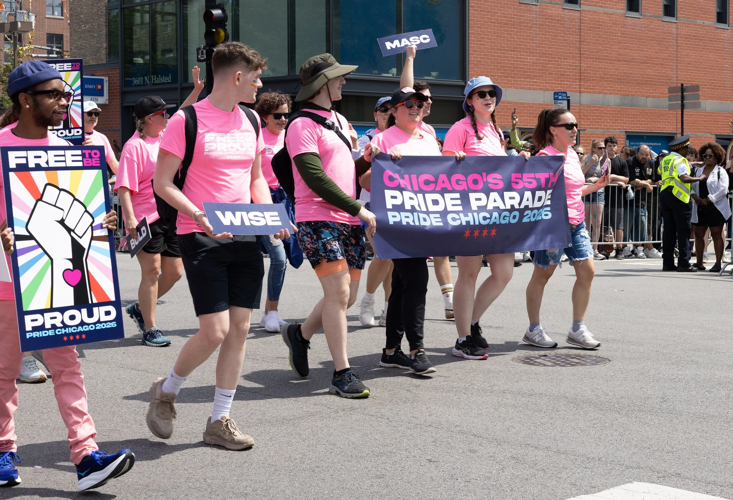 A group of diverse people marching in a pride parade, holding signs and banners that read "Chicago's 55th Pride Parade Pride Chicago 2026" and "Free to Be Proud." They are dressed in pink shirts and casual outfits, some wearing hats and sunglasses, w