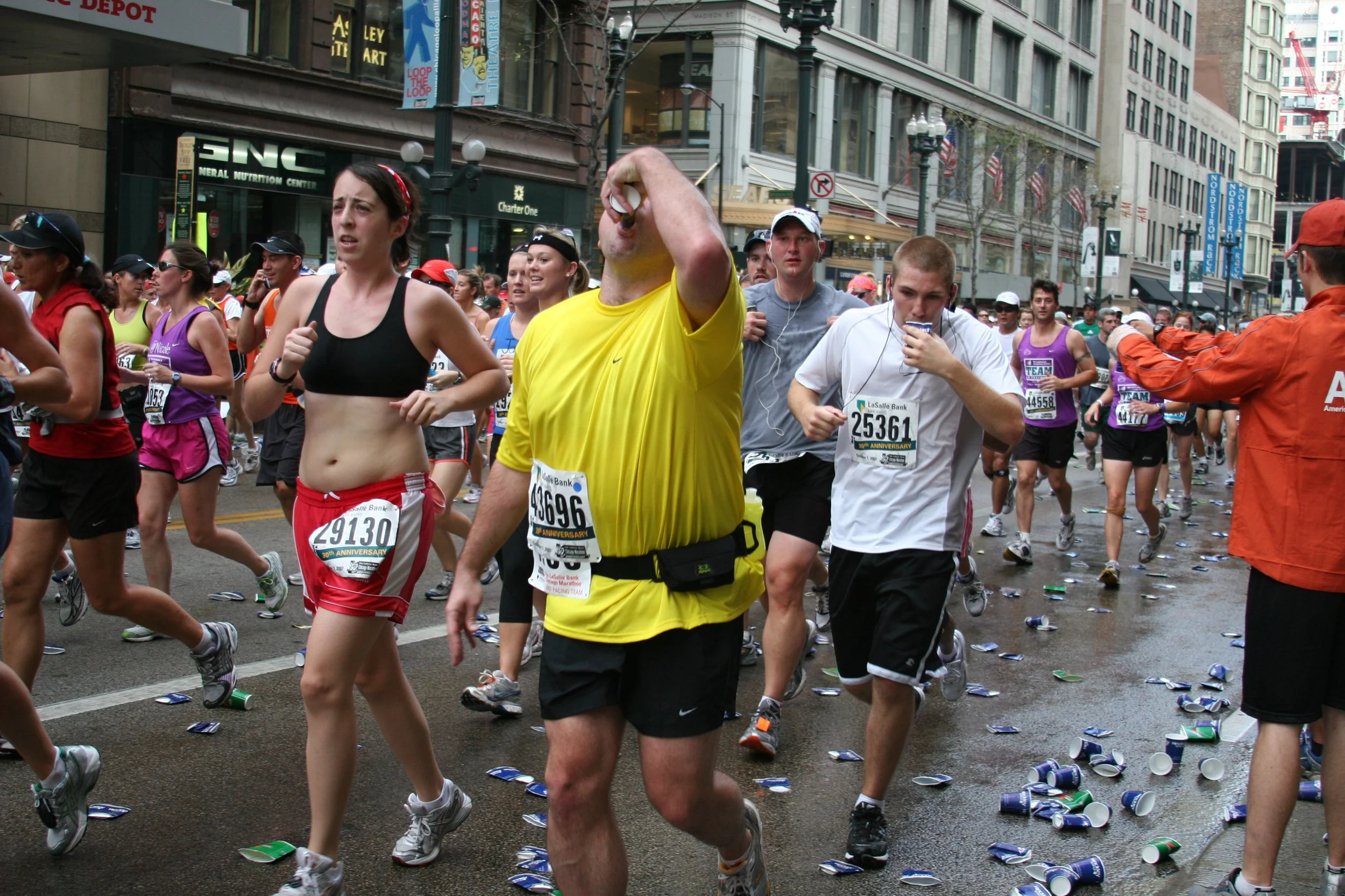 Crowd of marathon runners on city street with debris and trash, including paper cups, scattered on the road.