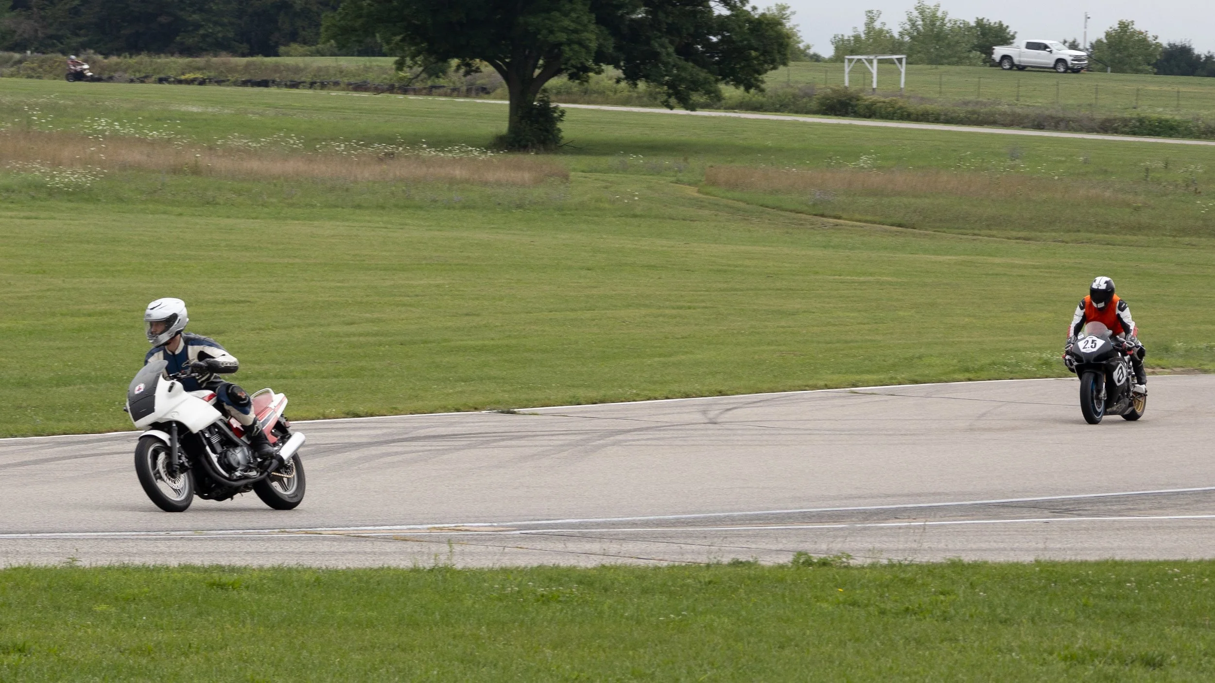 Two motorcyclists riding on a paved road through a grassy area with trees and a white truck in the background.