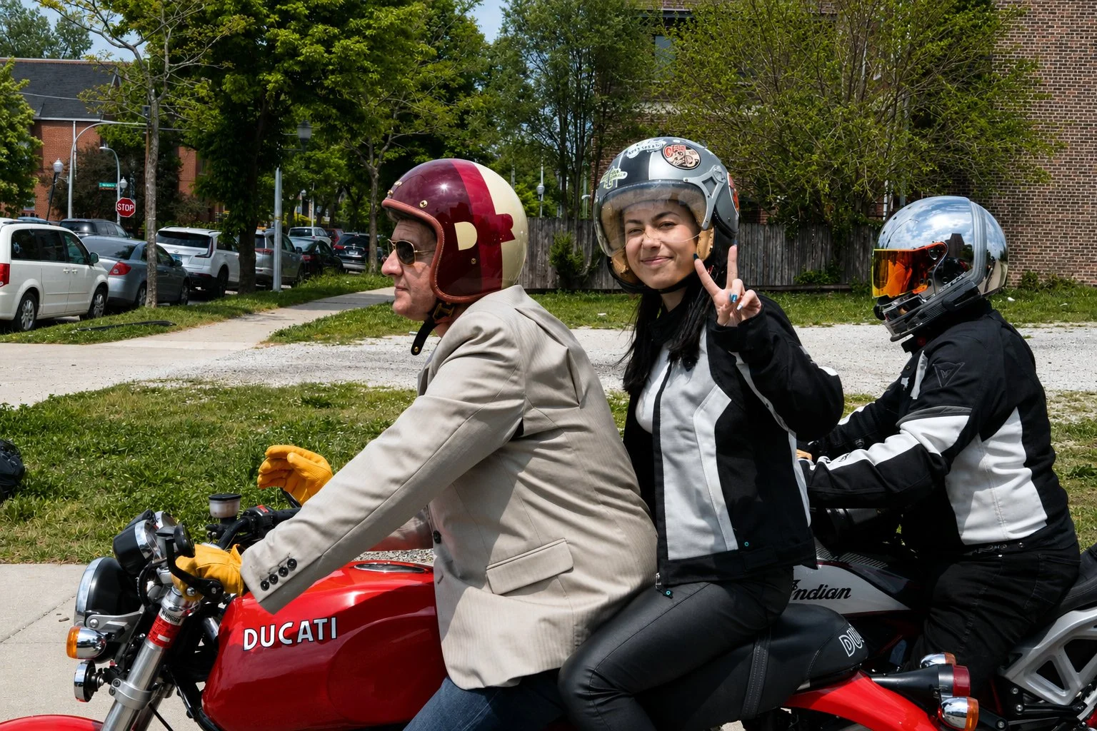 Three people riding a red Ducati motorcycle on a street, two wearing helmets and one woman making a peace sign and smiling.