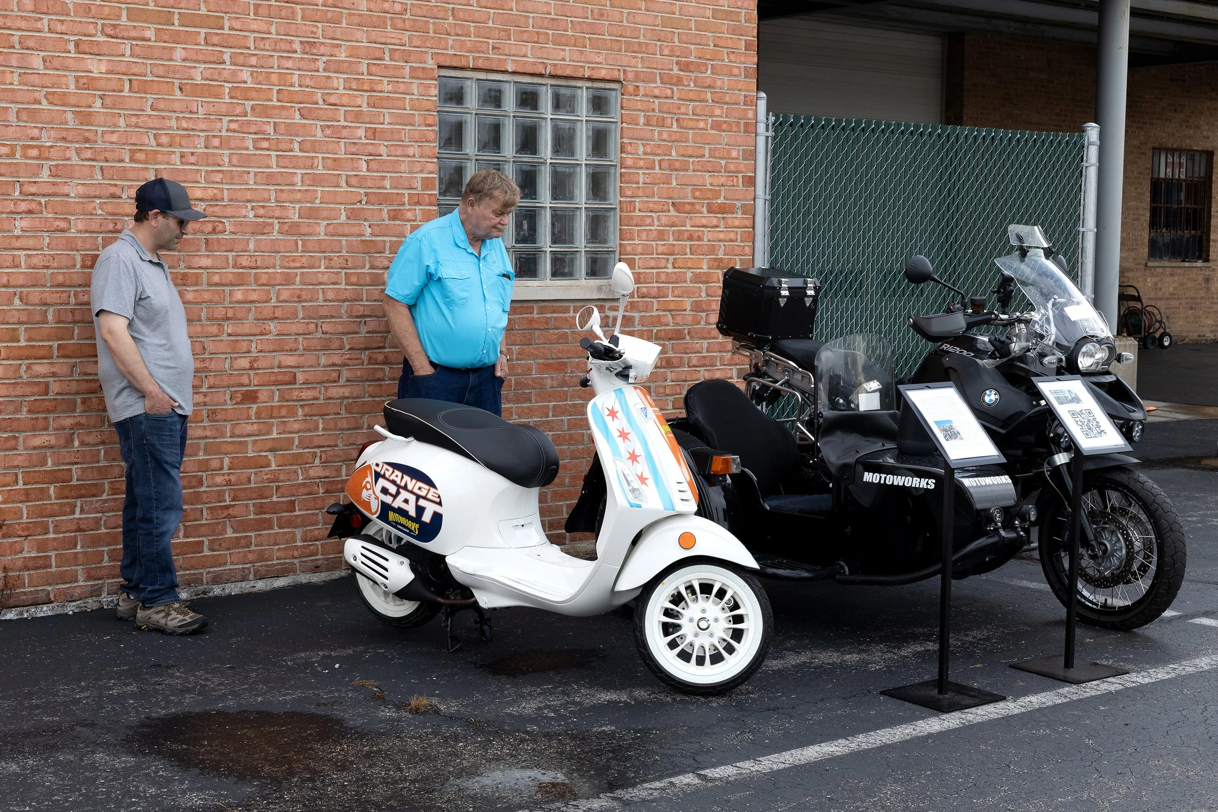 Two men standing next to a display of motorcycles and scooters outside a brick building, with a glass block window and green metal fence in the background.