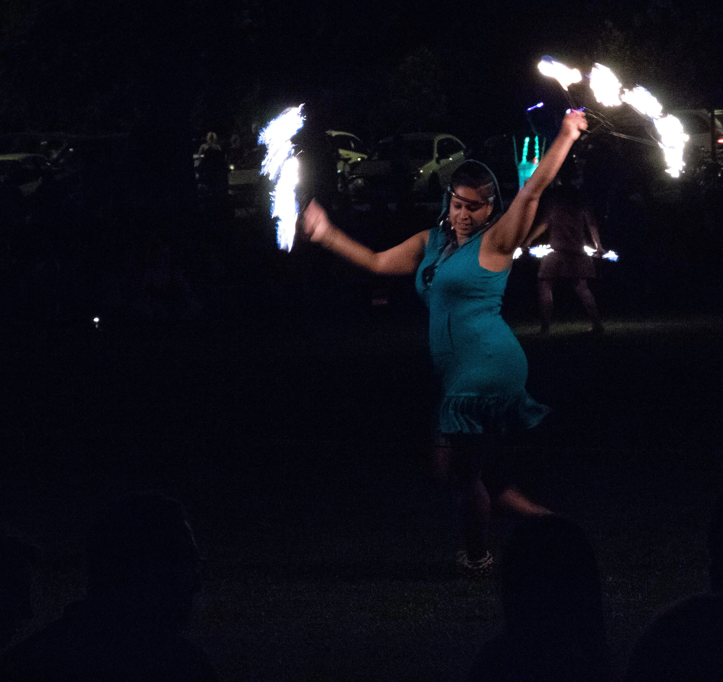 A person performing a fire spinning act at night, holding fire poi or fire staff, creating bright, fiery trails against a dark background, with vehicles parked nearby.