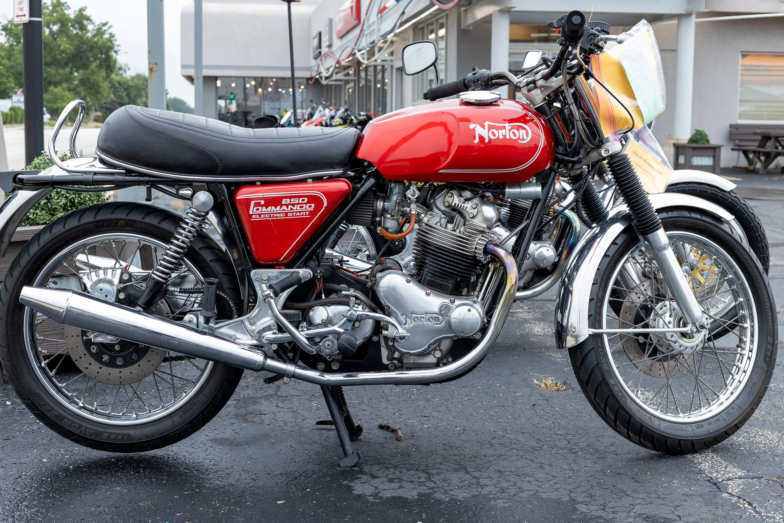 A red Norton motorcycle with a black seat, chrome exhaust, and detailed engine, parked on an asphalt lot outside a building with several other motorcycles in the background.