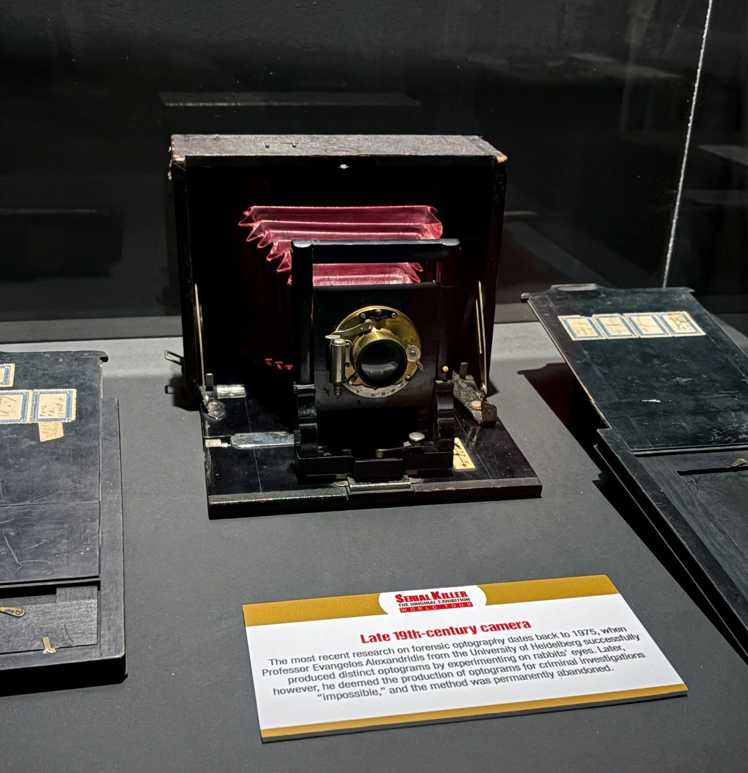 Close-up of an informational display about late 19th-century cameras with a partially visible red and black device and a printed sign explaining the history of forensic optography research.