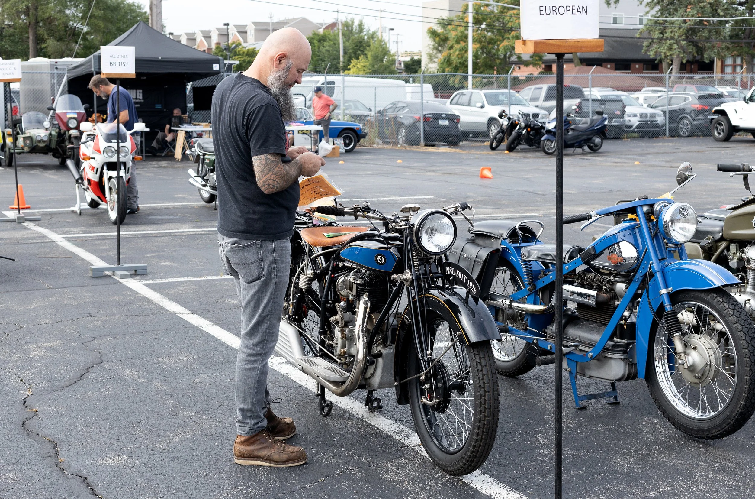A man with a beard and tattoos is examining a vintage motorcycle at an outdoor event. Other motorcycles and people are visible in the background, along with signs indicating different European countries.