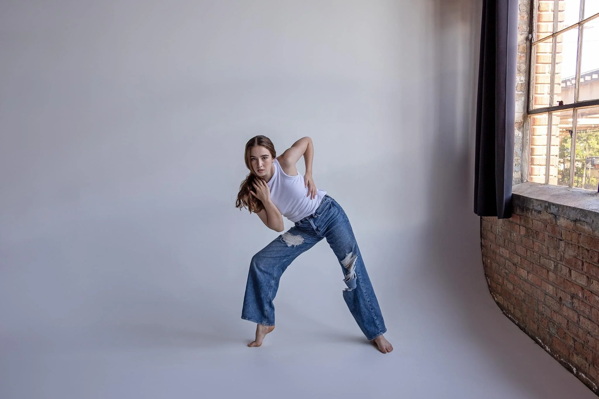 A woman in a white sleeveless shirt and ripped jeans is standing barefoot in a dance pose in front of a studio backdrop with a plain gray wall to her left and a large open window with brick walls outside to her right.