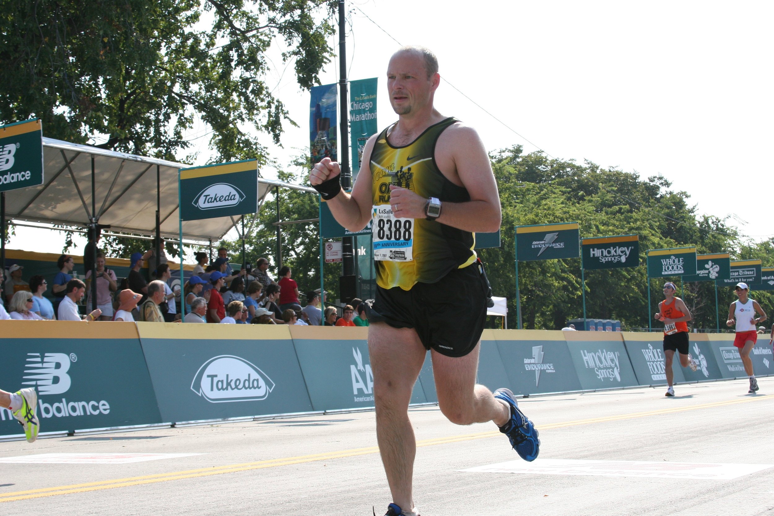 A man running in a marathon, wearing a yellow and black athletic shirt, black shorts, a watch, and a bib numbered 8388, with a crowd of spectators and other runners in the background during daytime.