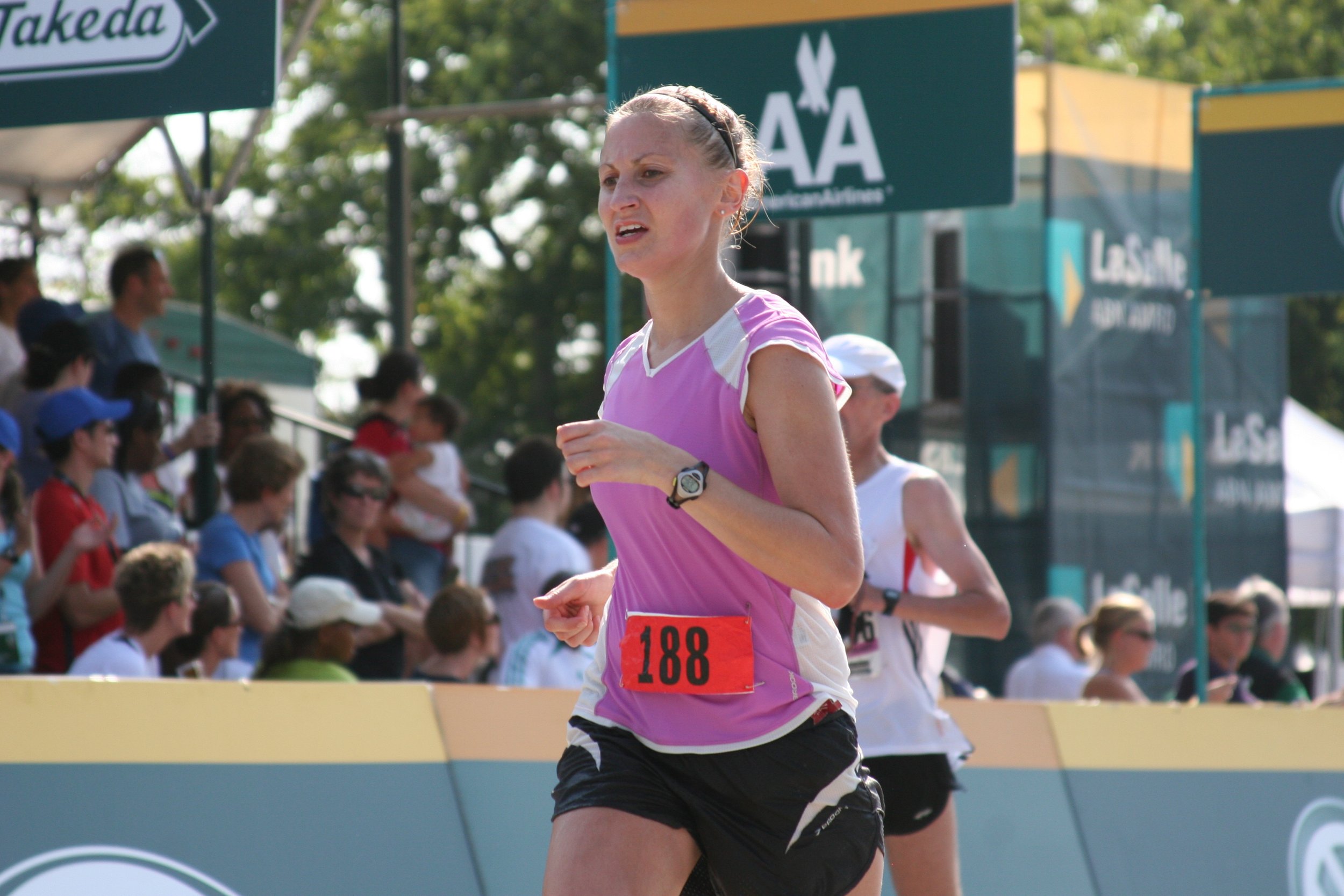 A female runner participating in a race, wearing a pink athletic outfit with bib number 188, running outdoors with a crowd of spectators in the background.
