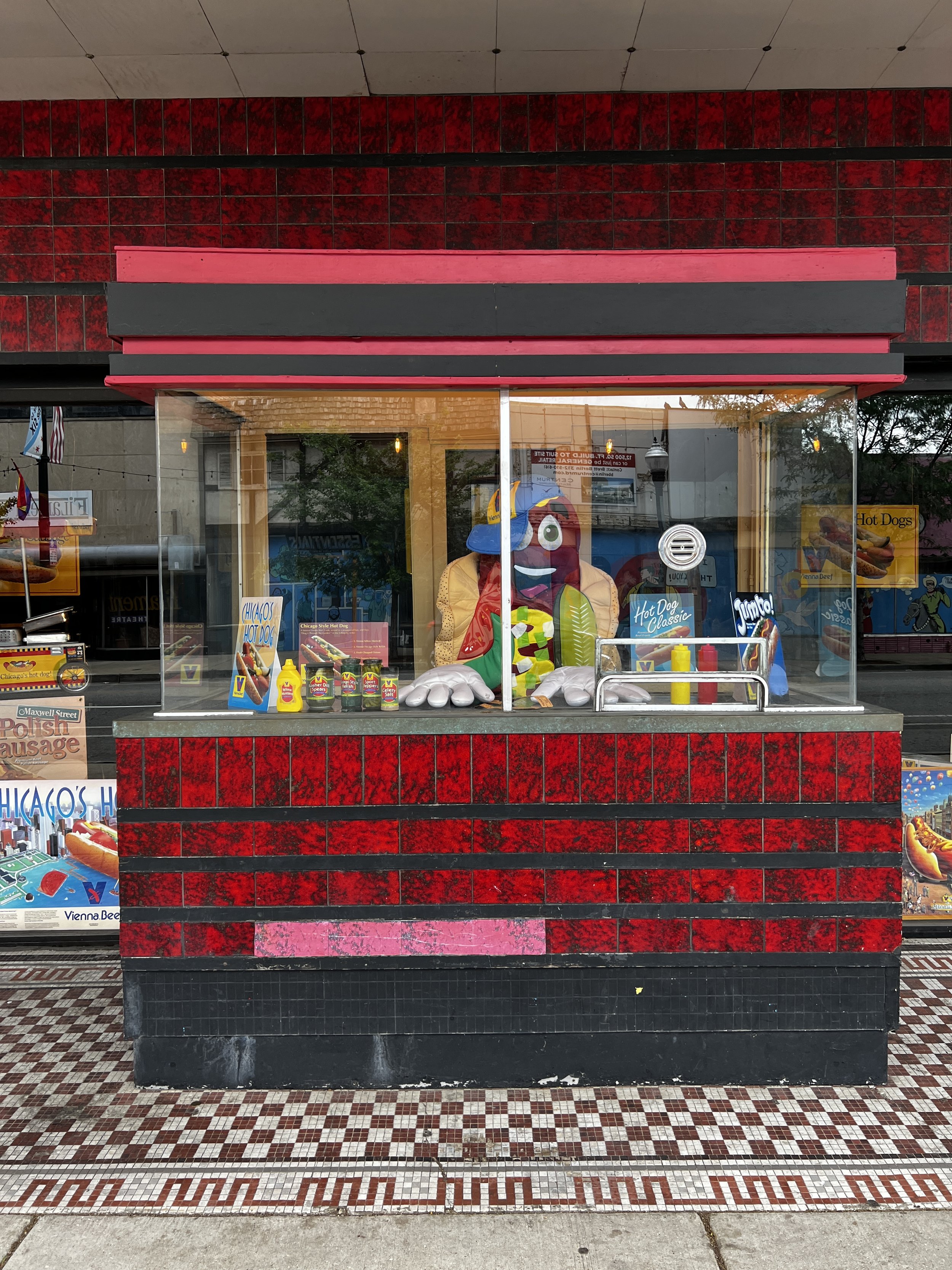 A storefront with red and black tiled wall, glass window, and a pizza mascot advertising hot dogs inside. Posters and signs are visible in the window.