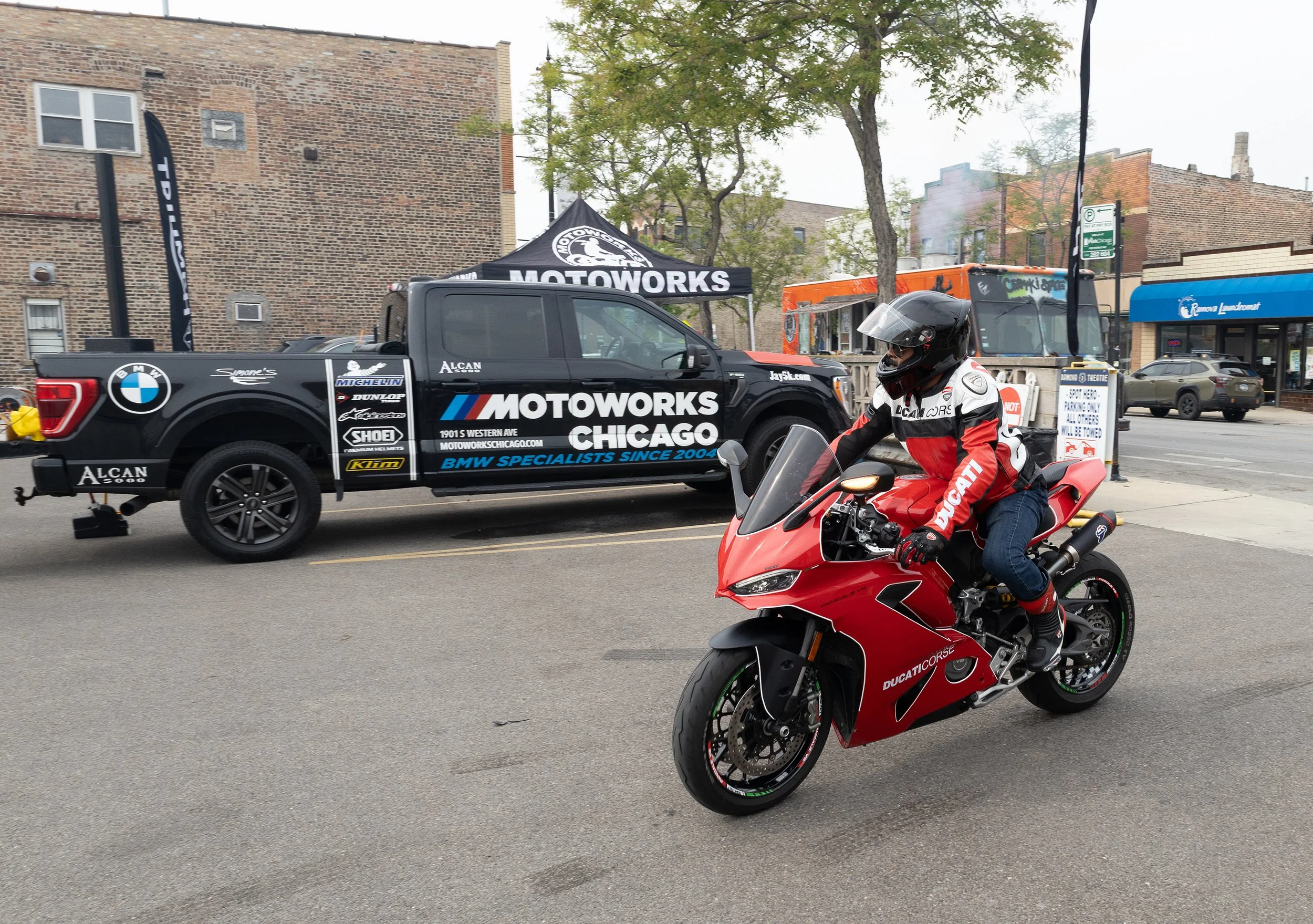 A street scene with a person in a red Ducati motorcycle riding in front of a BMW car with Motoworks Chicago advertisement and decals, a parked blue Crown Landmark store, and a billboard on a brick building.