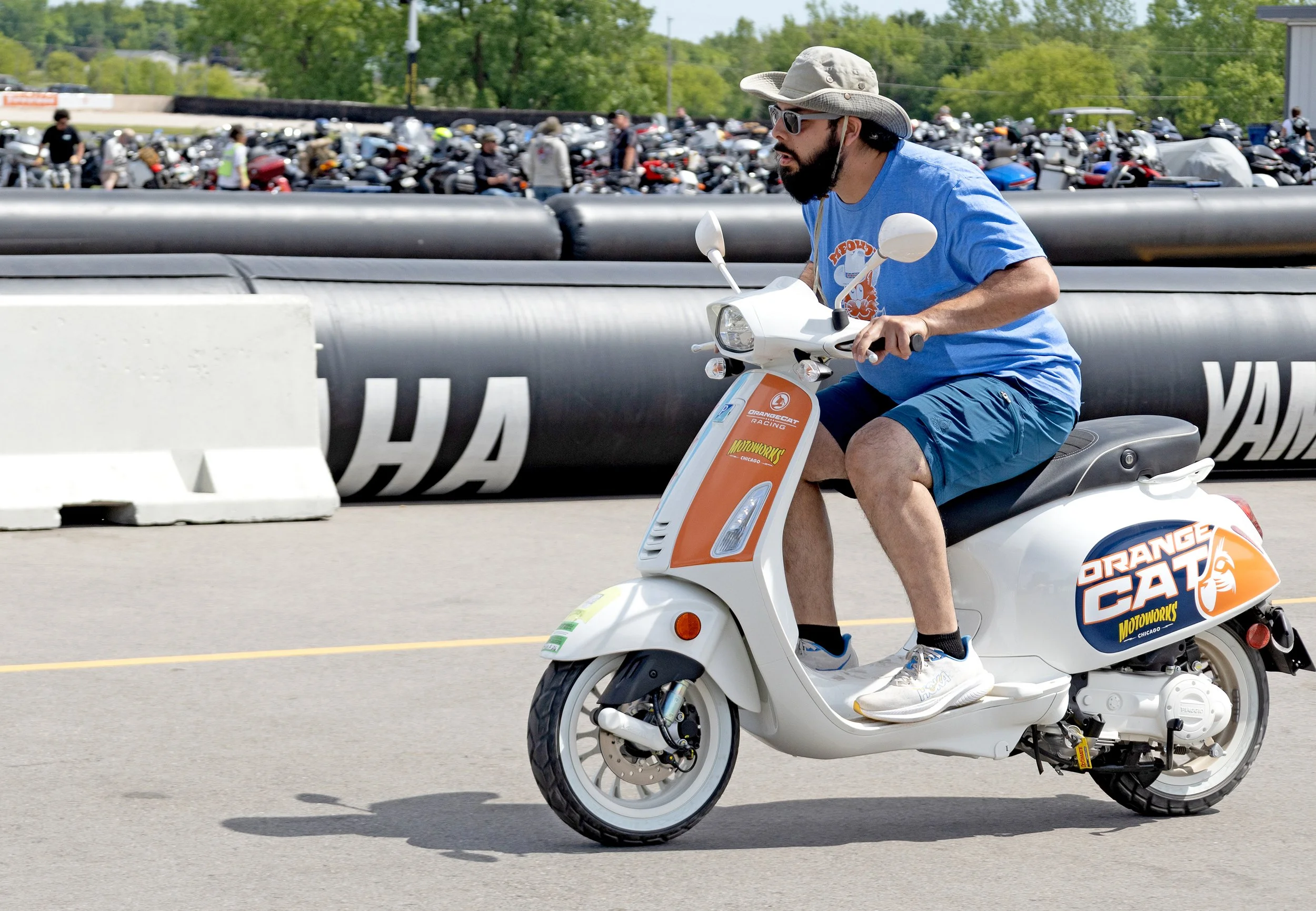 A man riding a white scooter with orange and blue branding, wearing a beige wide-brimmed hat, sunglasses, a blue t-shirt, and shorts, with a busy parking lot filled with motorcycles in the background.