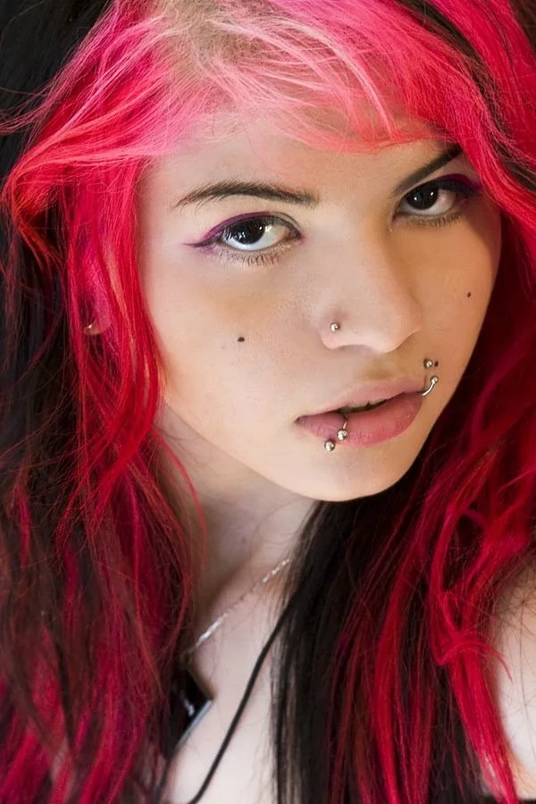 Close-up of a young woman with vibrant red and black hair, multiple facial piercings, and pink eye makeup.