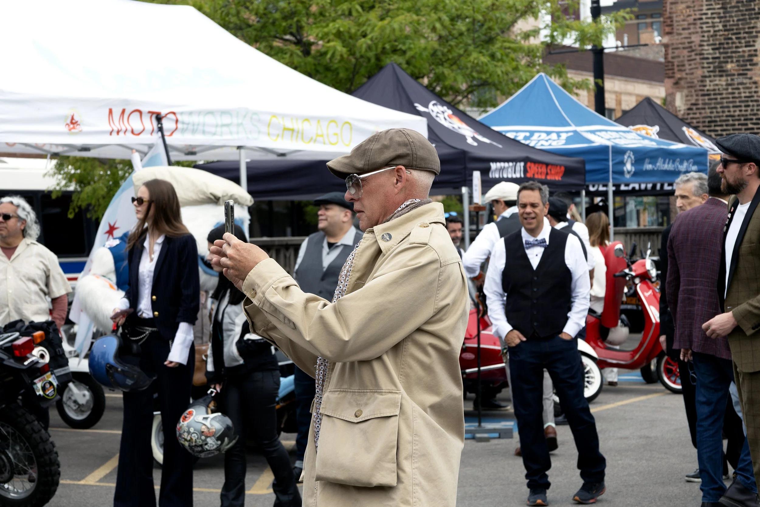 A man in a beige trench coat and brown cap taking a photo at an outdoor event with tents and people in vintage attire, some with motorcycles and scooters, in a city street.