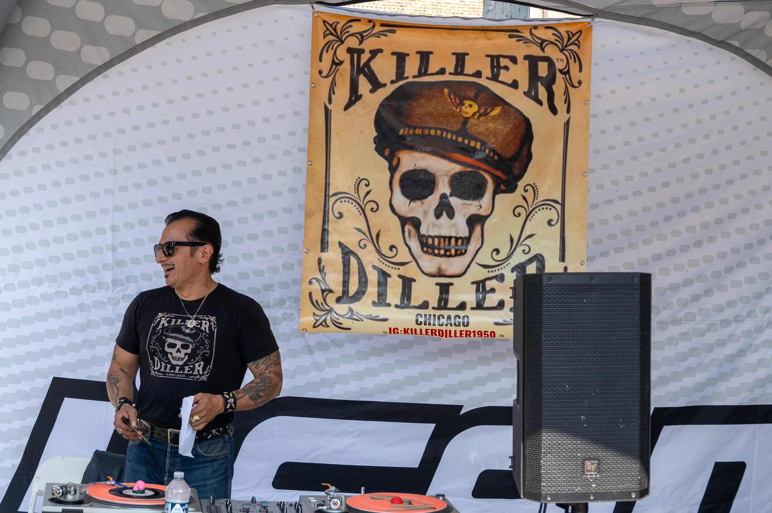 A man wearing sunglasses, a black T-shirt with a skull and 'KILLER DILLER' logo, and tattoos on his arms, is smiling while standing at a DJ booth with turntables and equipment. Behind him, a large banner displays the 'KILLER DILLER' logo with a skull
