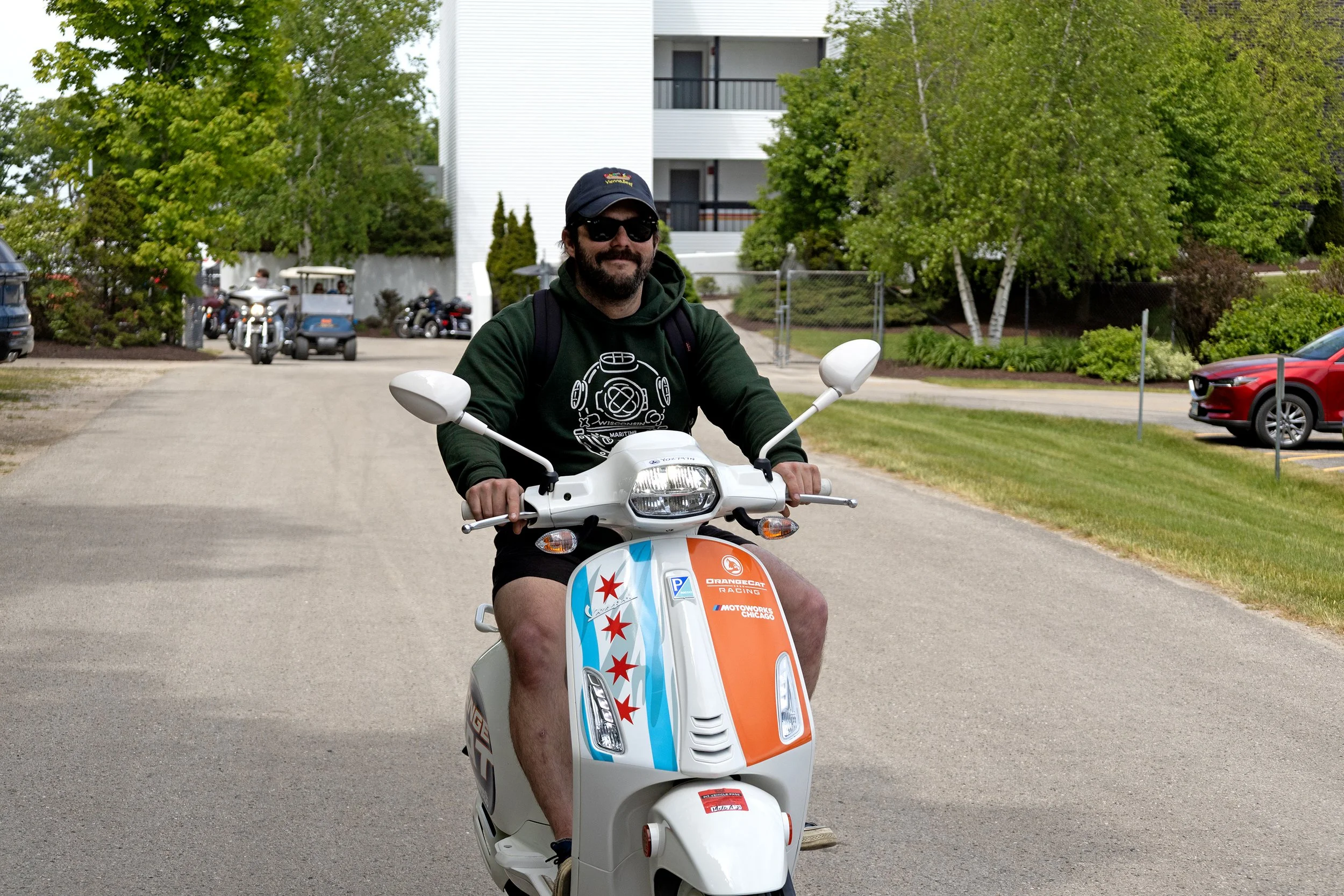 A man with a beard, sunglasses, and a dark hoodie riding a white scooter decorated with Chicago Cubs colors and logos, on a road with trees and apartments in the background.