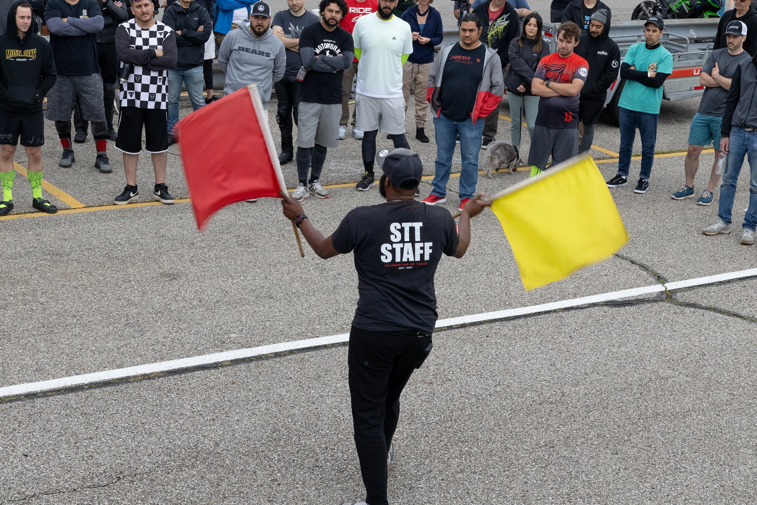 A group of people standing in a parking lot, some with their arms crossed, watching a person waving yellow and red flags. The person with flags is wearing a black shirt with white text on the back that says 'STT STAFF' and a black cap. The group appe
