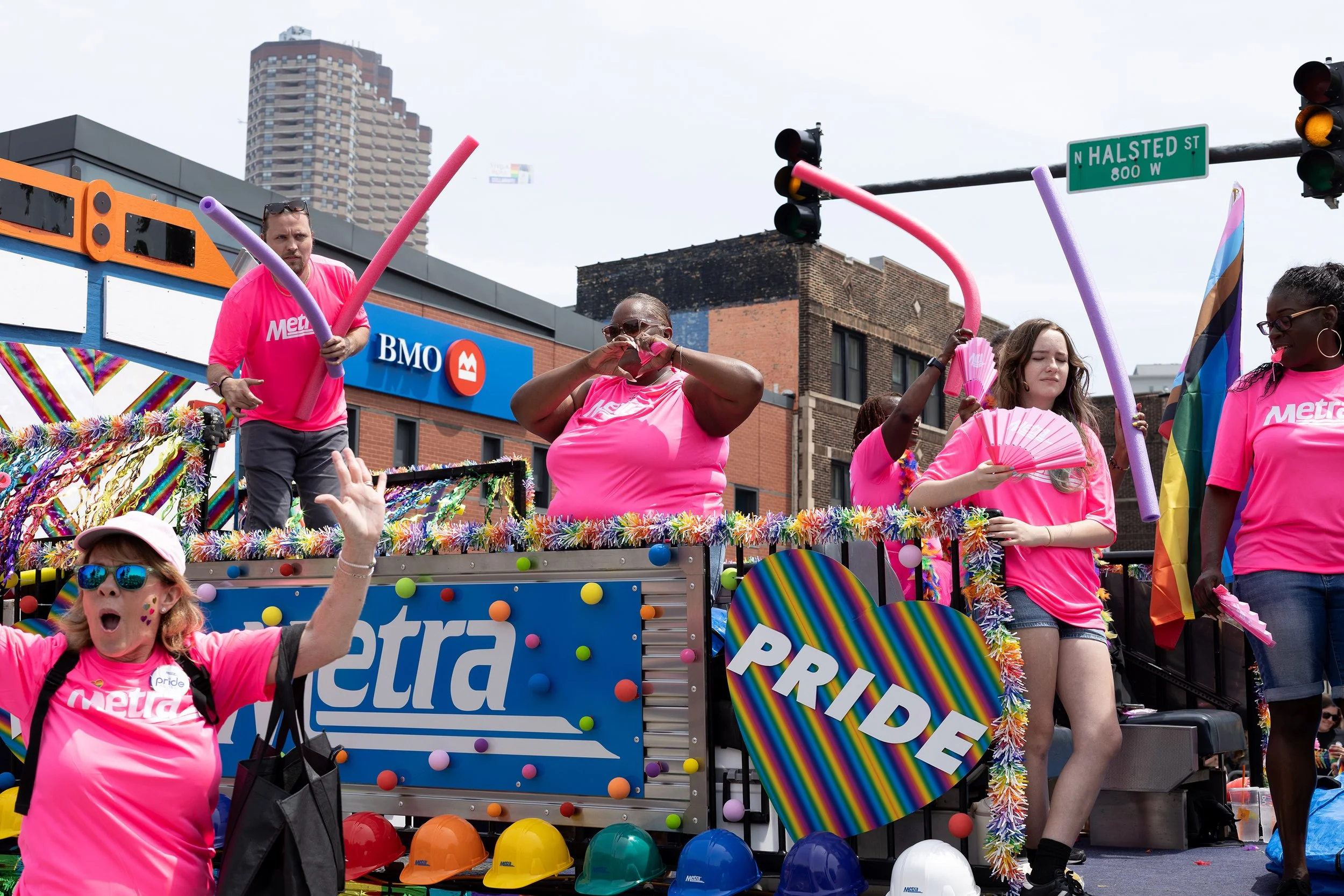 People participating in a pride parade, wearing pink shirts, some with rainbow accessories, on a decorated float with rainbow elements and signs, including one that reads 'PRIDE.' A rainbow flag is visible, and participants are celebrating.