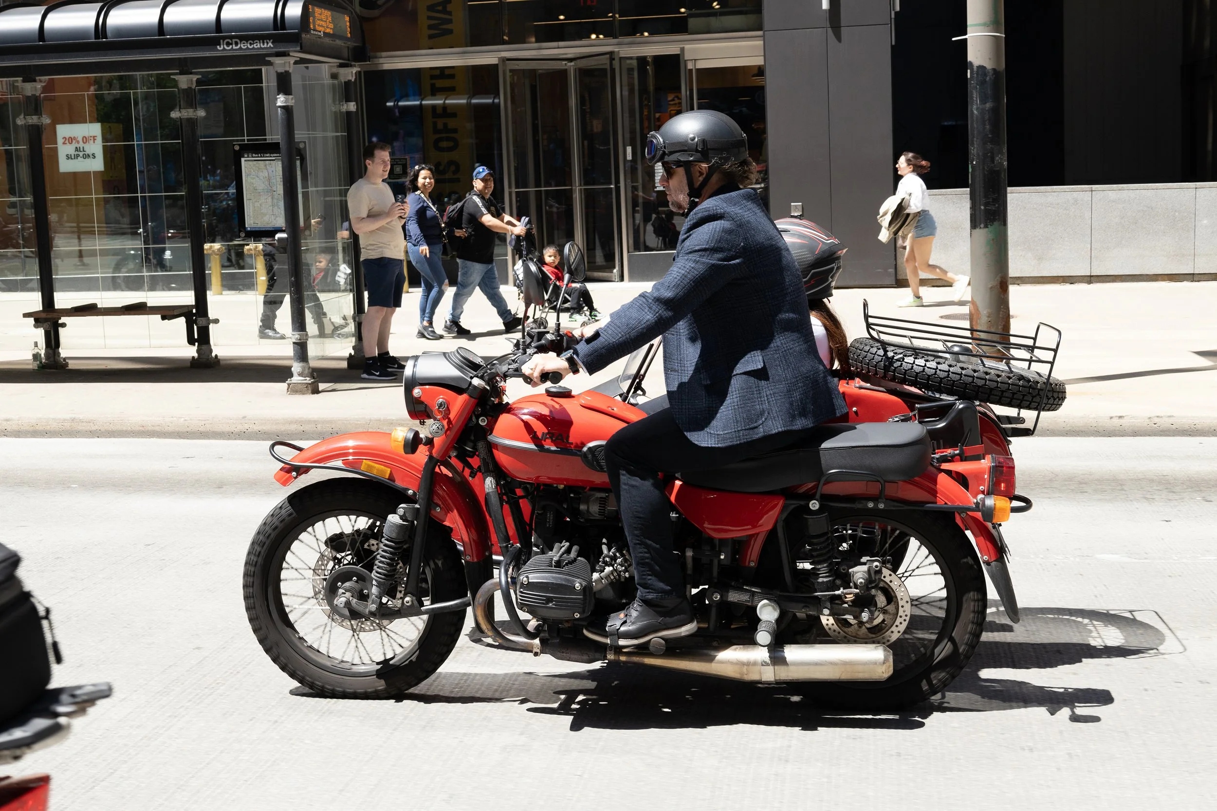 Man riding a red motorcycle on the city street with pedestrians in the background.