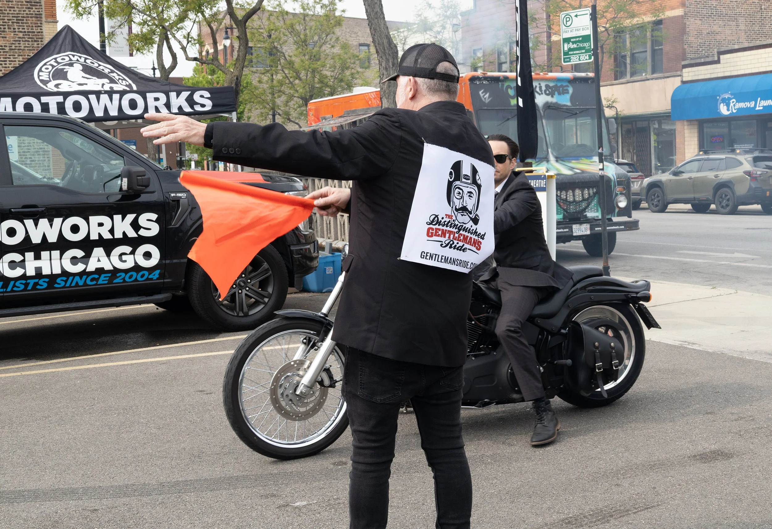 A man on a motorcycle and a woman standing on the street during the day. The man is wearing black clothing and sunglasses, riding a black motorcycle, while the woman in a black jacket has a sign with a cartoon logo on her back. The street has parked 