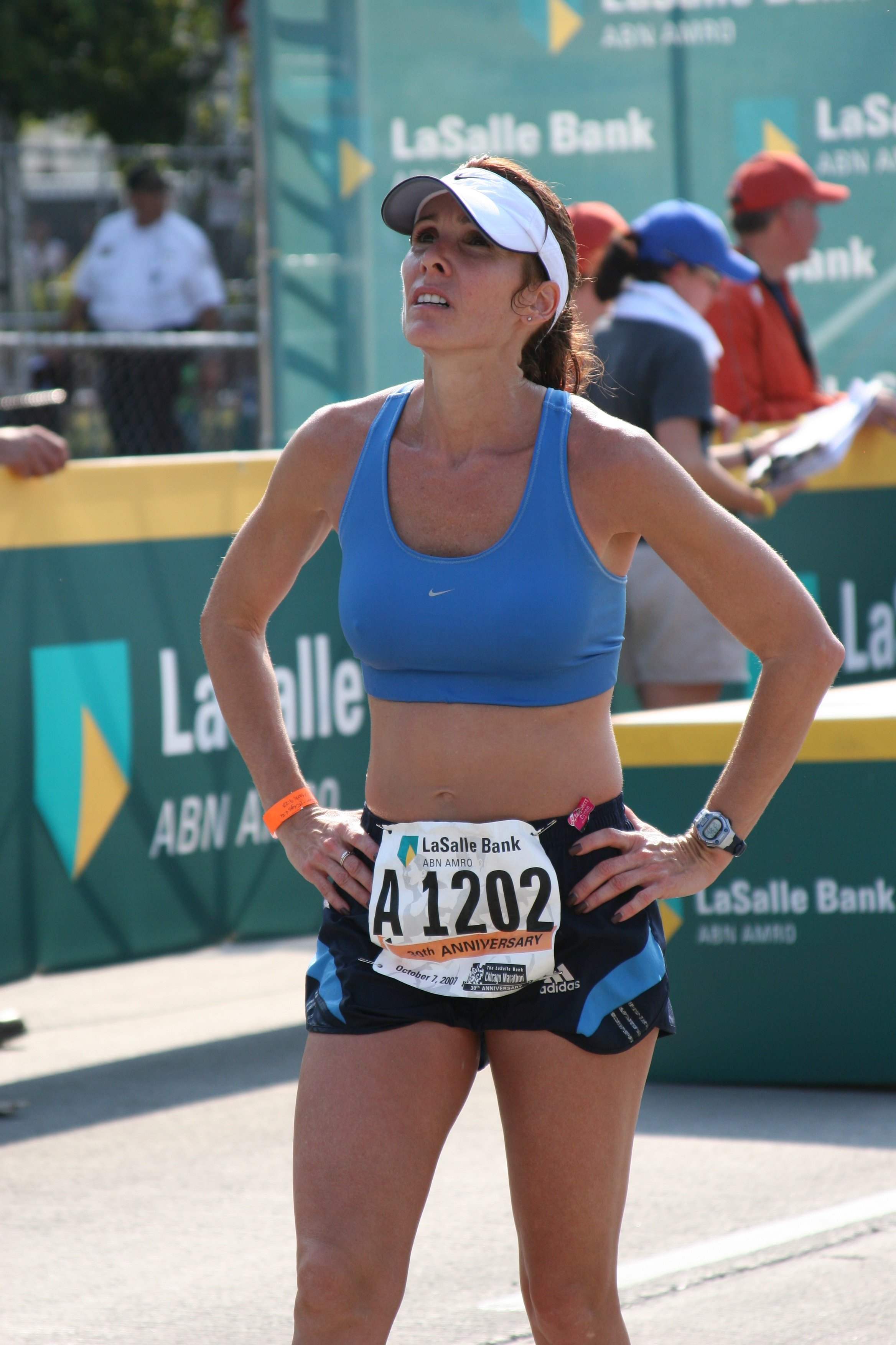 A female marathon runner in a blue sports bra and black shorts, standing with her hands on her hips during a race. She is wearing a white visor and a watch. Race bib number A1202 is visible. The background features a green banner with 'LaSalle Bank' 