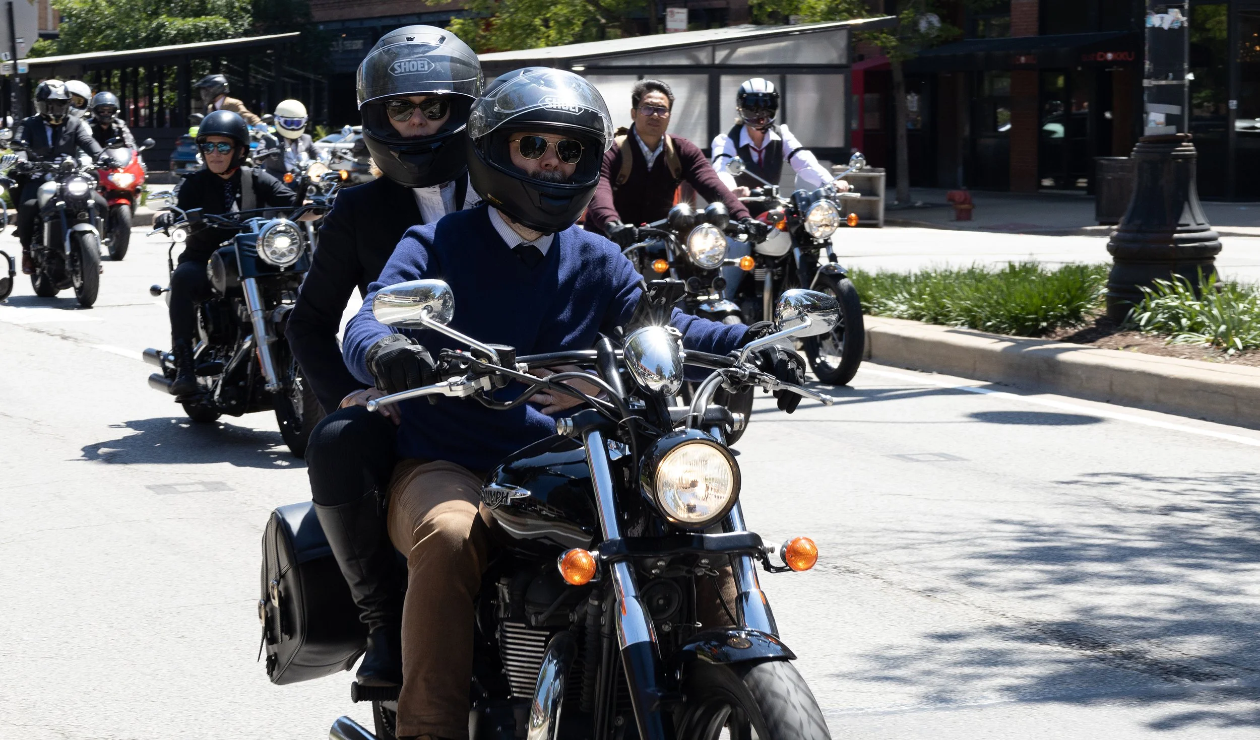 A group of people riding motorcycles on a city street on a sunny day, with some wearing helmets and sunglasses.