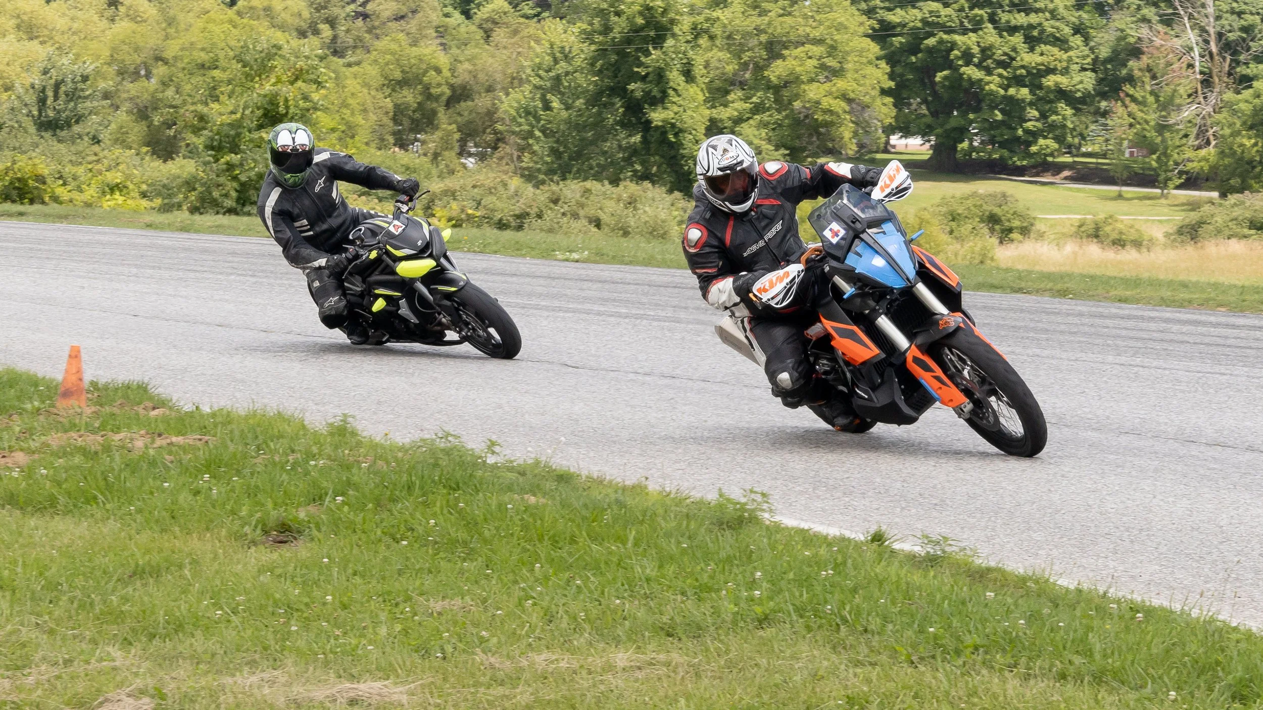Two motorcyclists wearing protective gear leaning into a turn on a paved road surrounded by green trees and grass.
