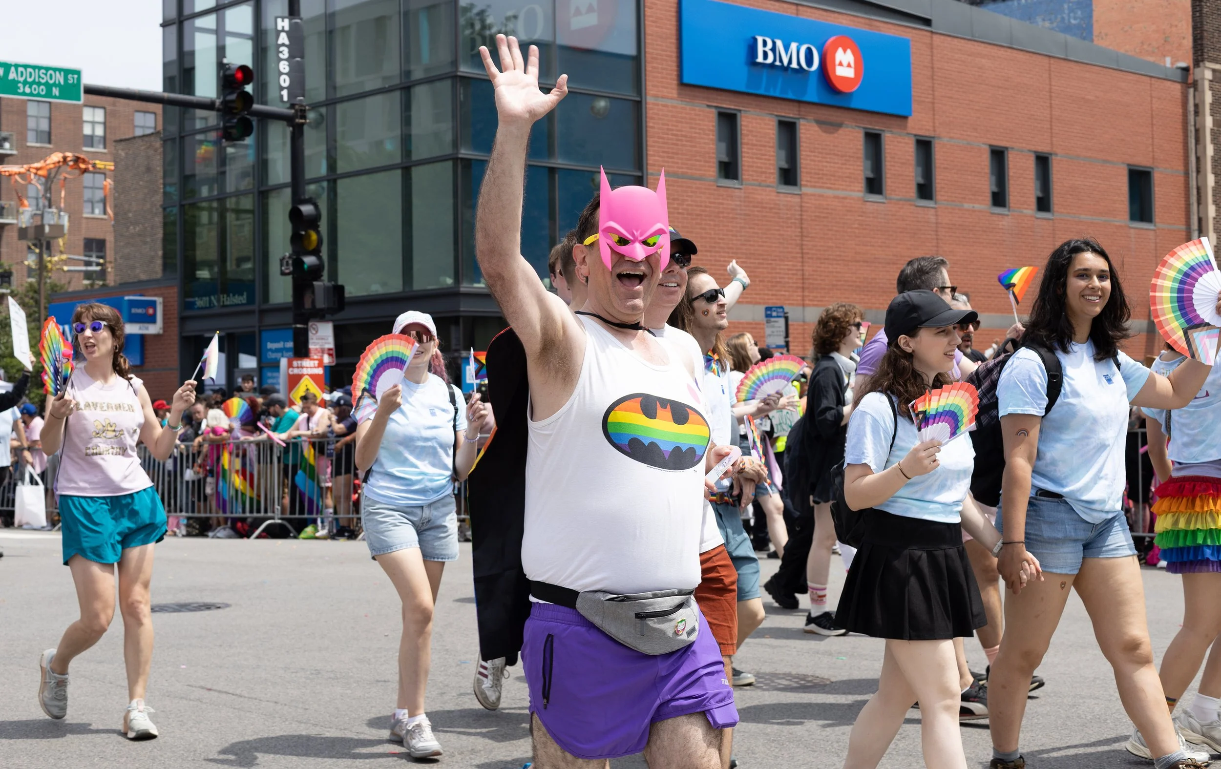 People participating in a pride parade, wearing rainbow flags and celebrating, with a man in the foreground wearing a pink Batman mask and a tank top with a rainbow Batman logo, in front of a city building with a BMO sign.