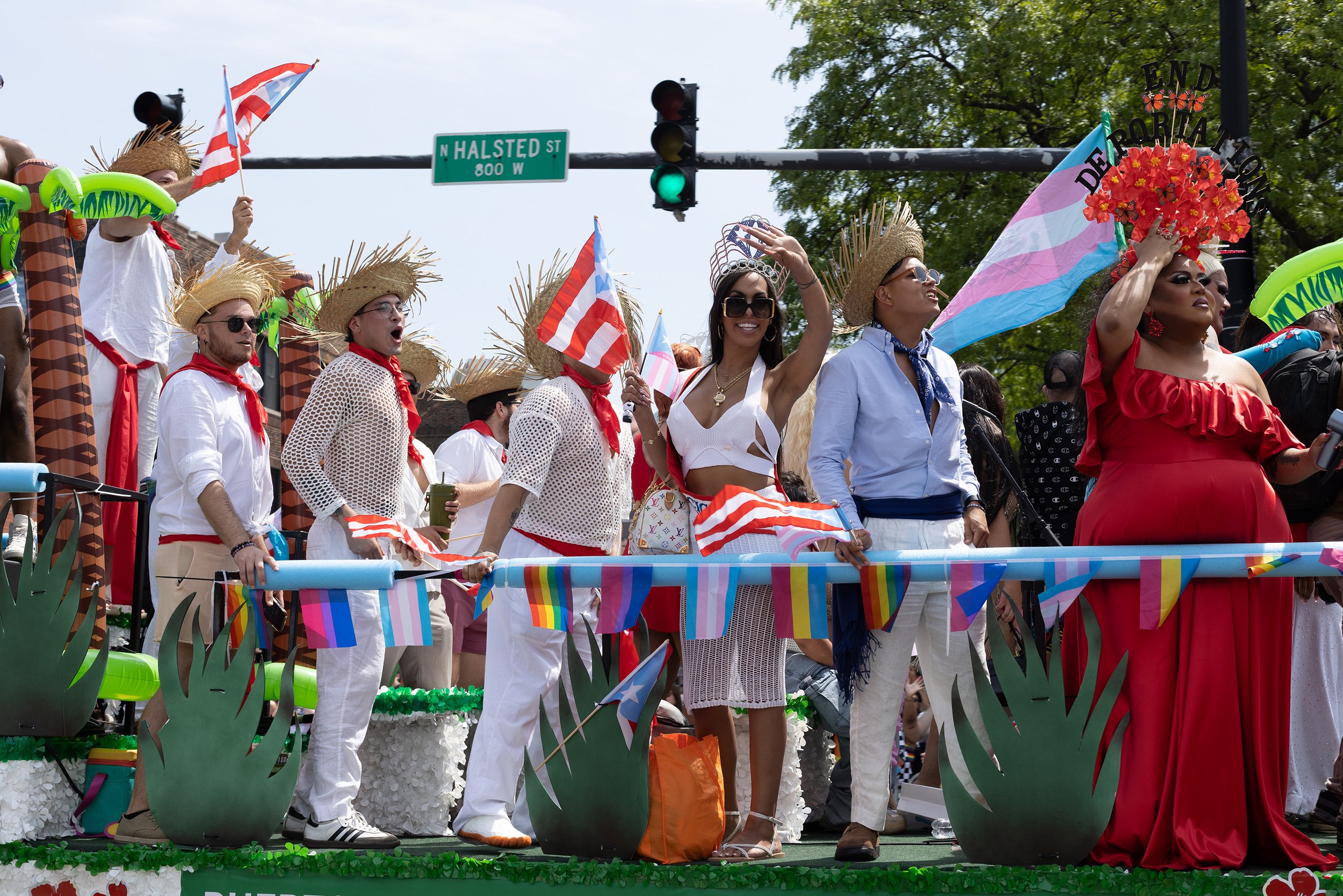 People dressed in colorful, festive costumes riding on a parade float decorated with rainbow flags and tropical-themed decorations during a pride parade on a city street.
