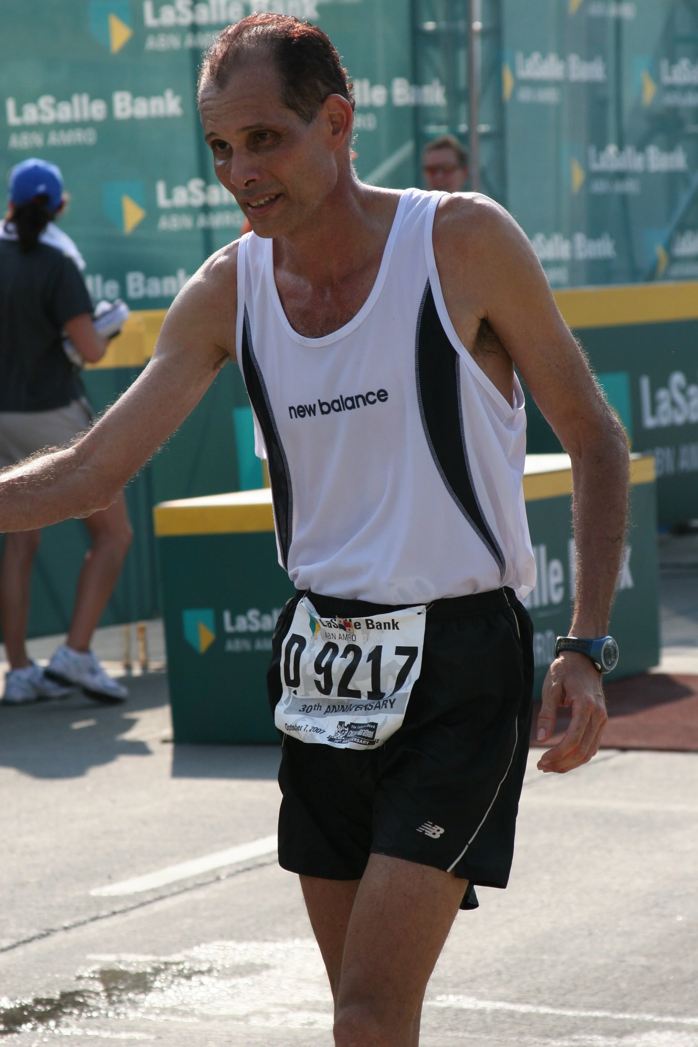 A male marathon runner wearing a white New Balance tank top and black shorts, with a bib number 09217, seen during a race in Los Angeles, with a green background featuring LaSalle Bank signage.