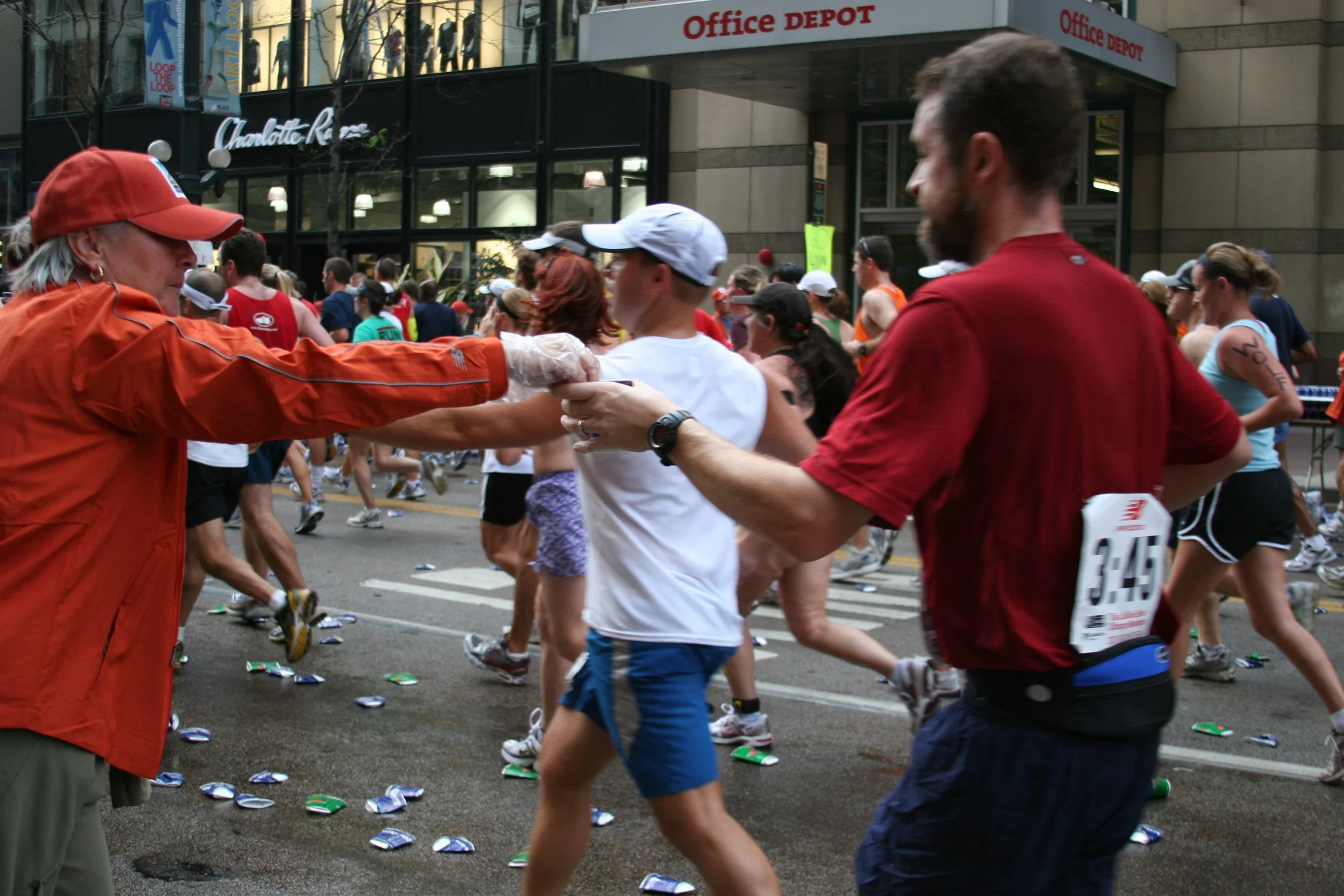 People participating in a running event on city streets, with a crowd and buildings, including Office Depot and Charlotte Russe signs, in the background. Cans of soda are scattered on the ground.