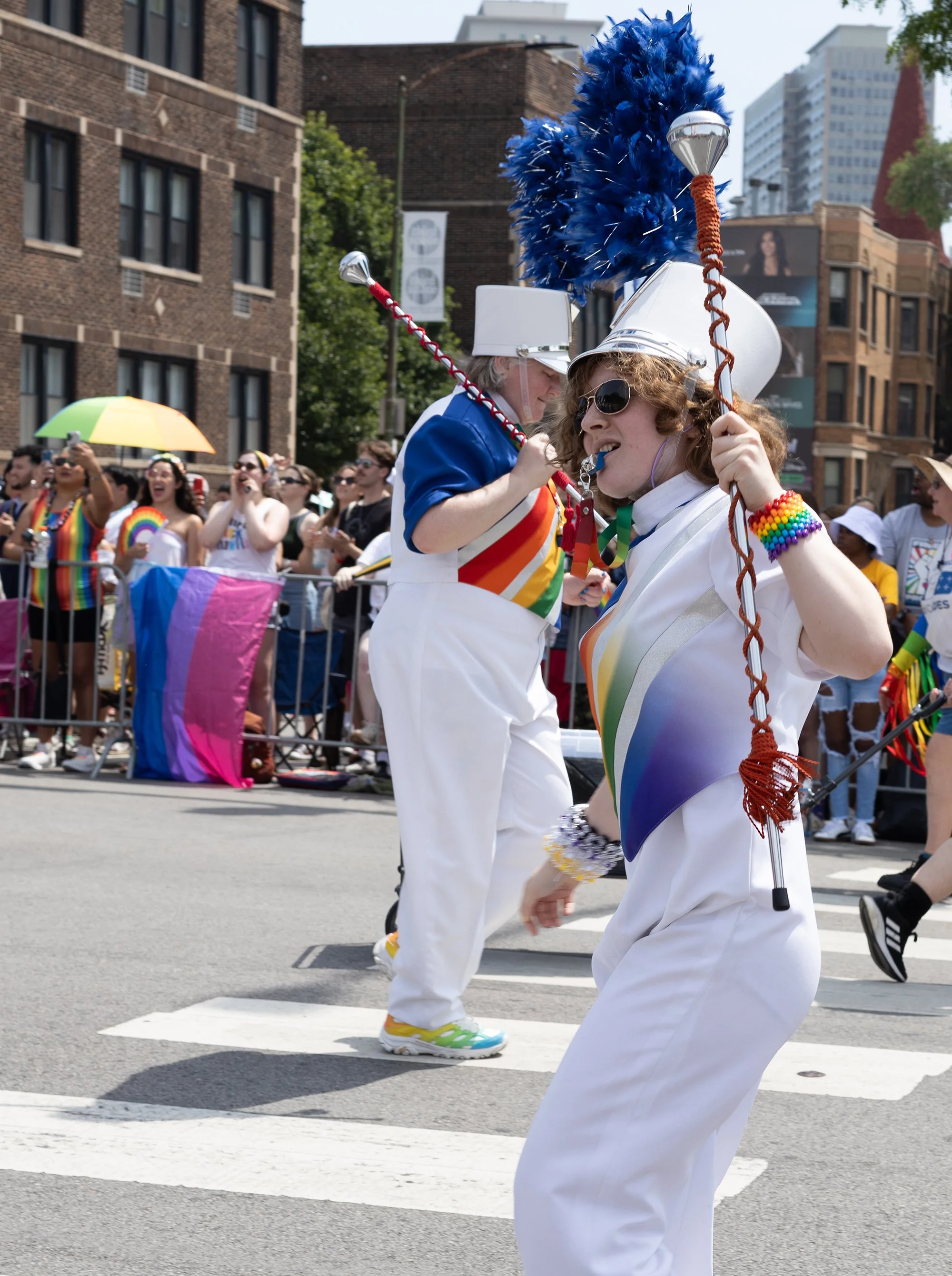 Two people dressed in white with rainbow accessories are dancing at a Pride parade. They are wearing white hats with blue and orange feathers, and rainbow flags are visible in the background. One person is wearing sunglasses and speaking into a whist