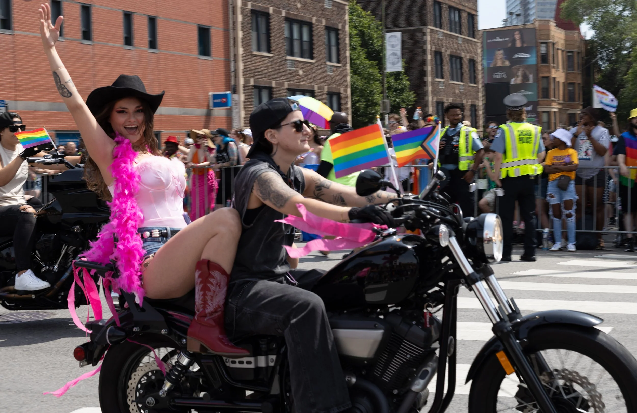 A woman riding a motorcycle during a pride parade, with people holding rainbow flags in the background. She is wearing a black hat, pink feather boa, and smiling.