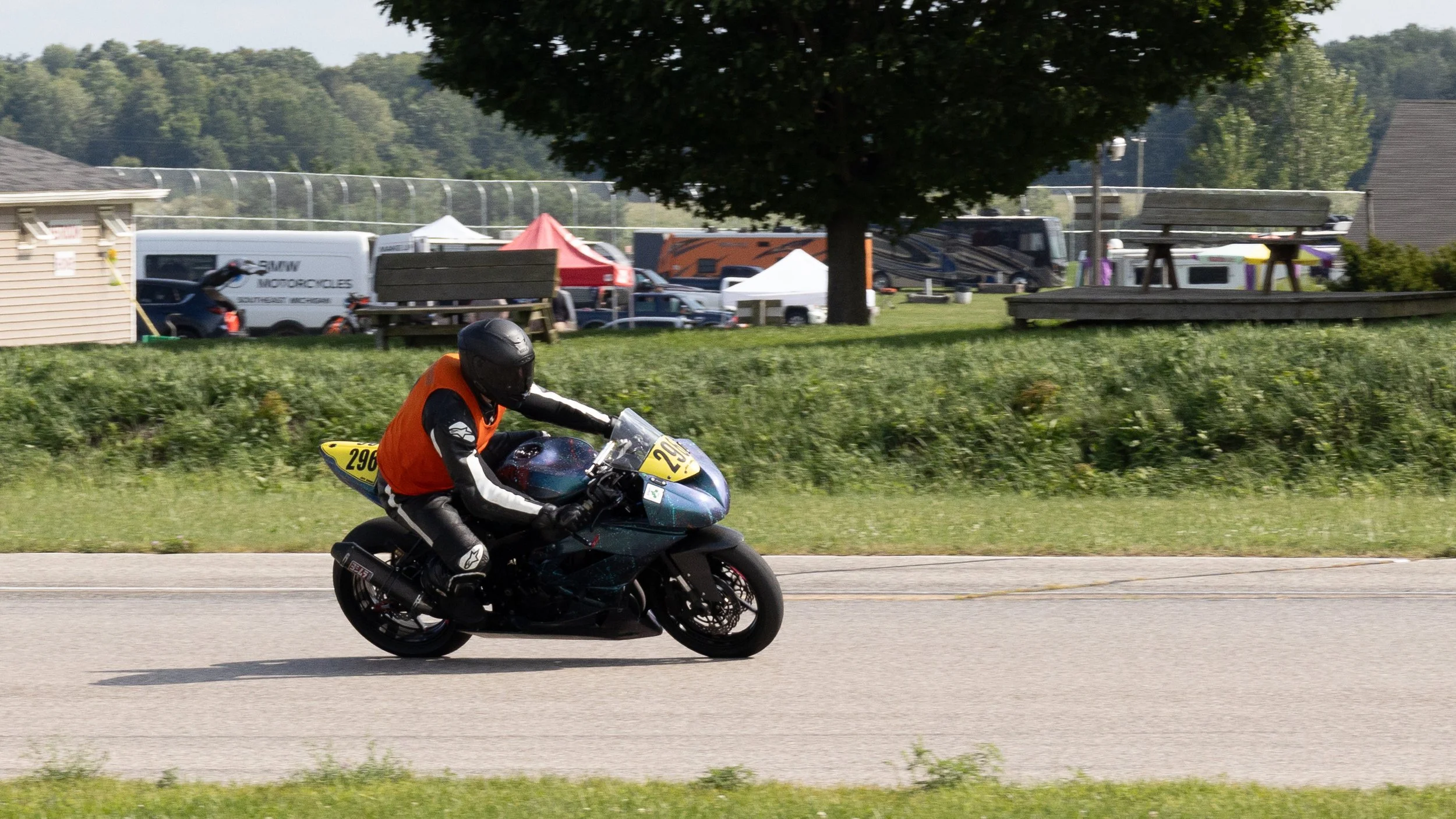 Motorcycle racer wearing an orange vest and black helmet speeding along a road with a blurred background of tents, vehicles, and greenery.