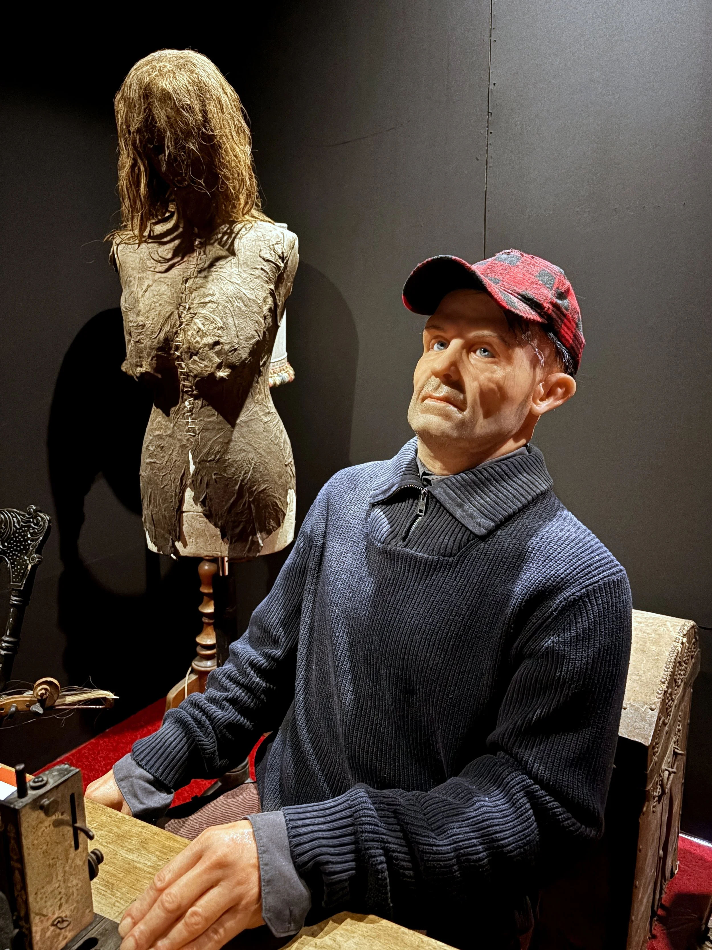 An older man with a red and gray plaid hat, sitting at a table with showing a focused expression. Behind him is a mannequin with a long, curly blonde wig against a dark background.