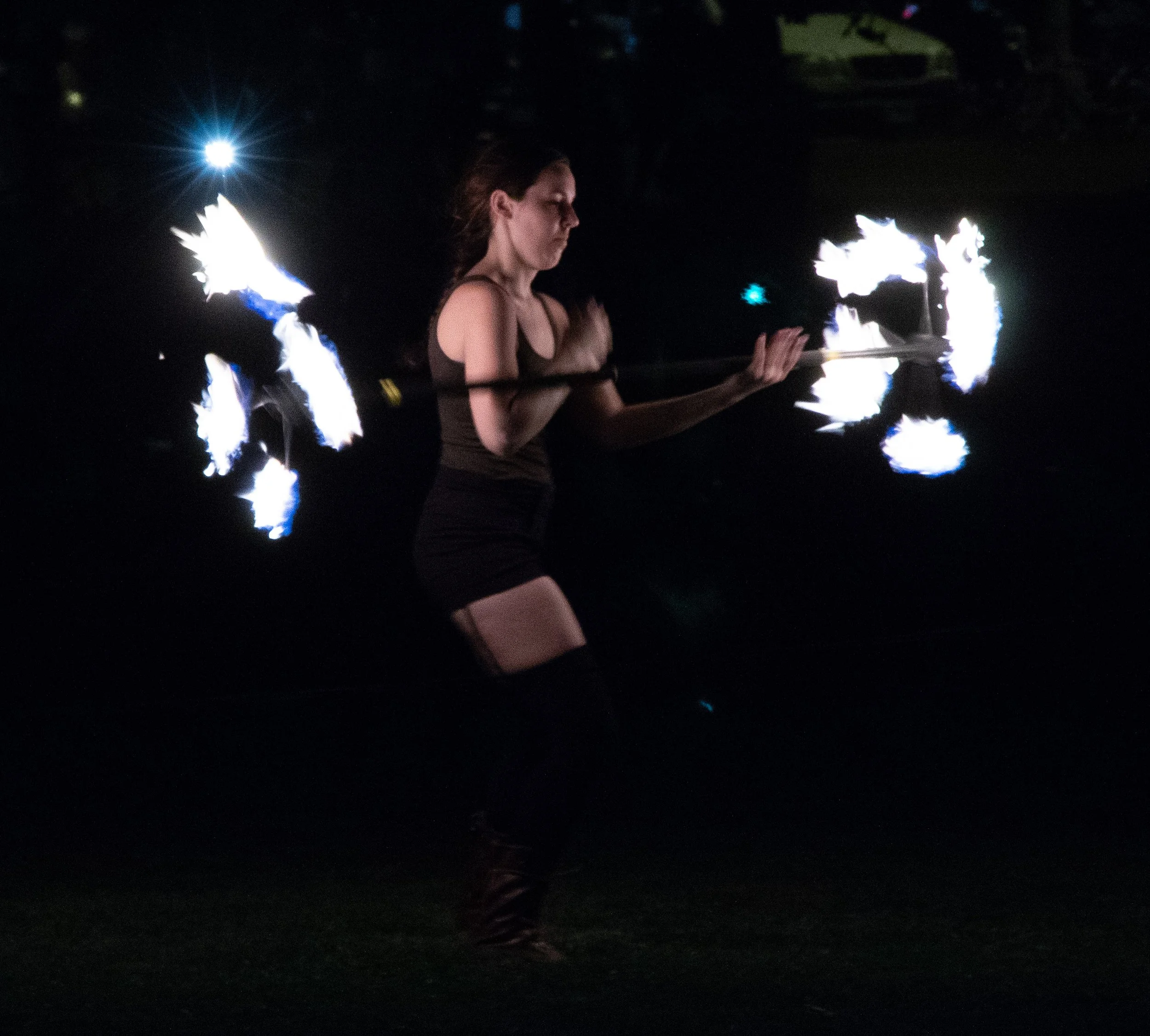A woman holding a flaming torch in each hand at night, with flames reflecting on her face and body.