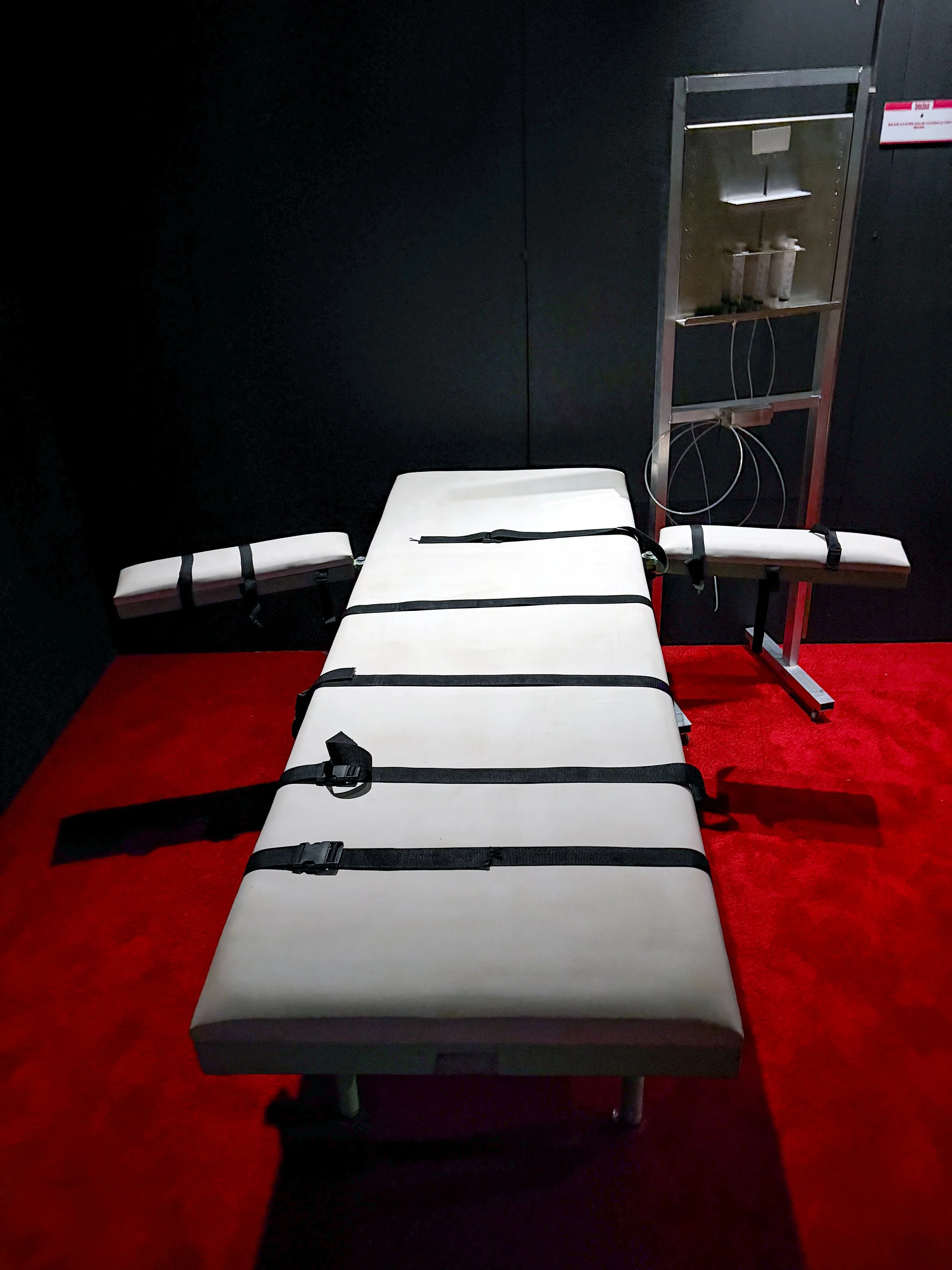 A medical examination table with a white cushion, straps, and a black belt, placed on a red carpeted floor against a black wall, with a metal medical lamp and equipment stand to the side.