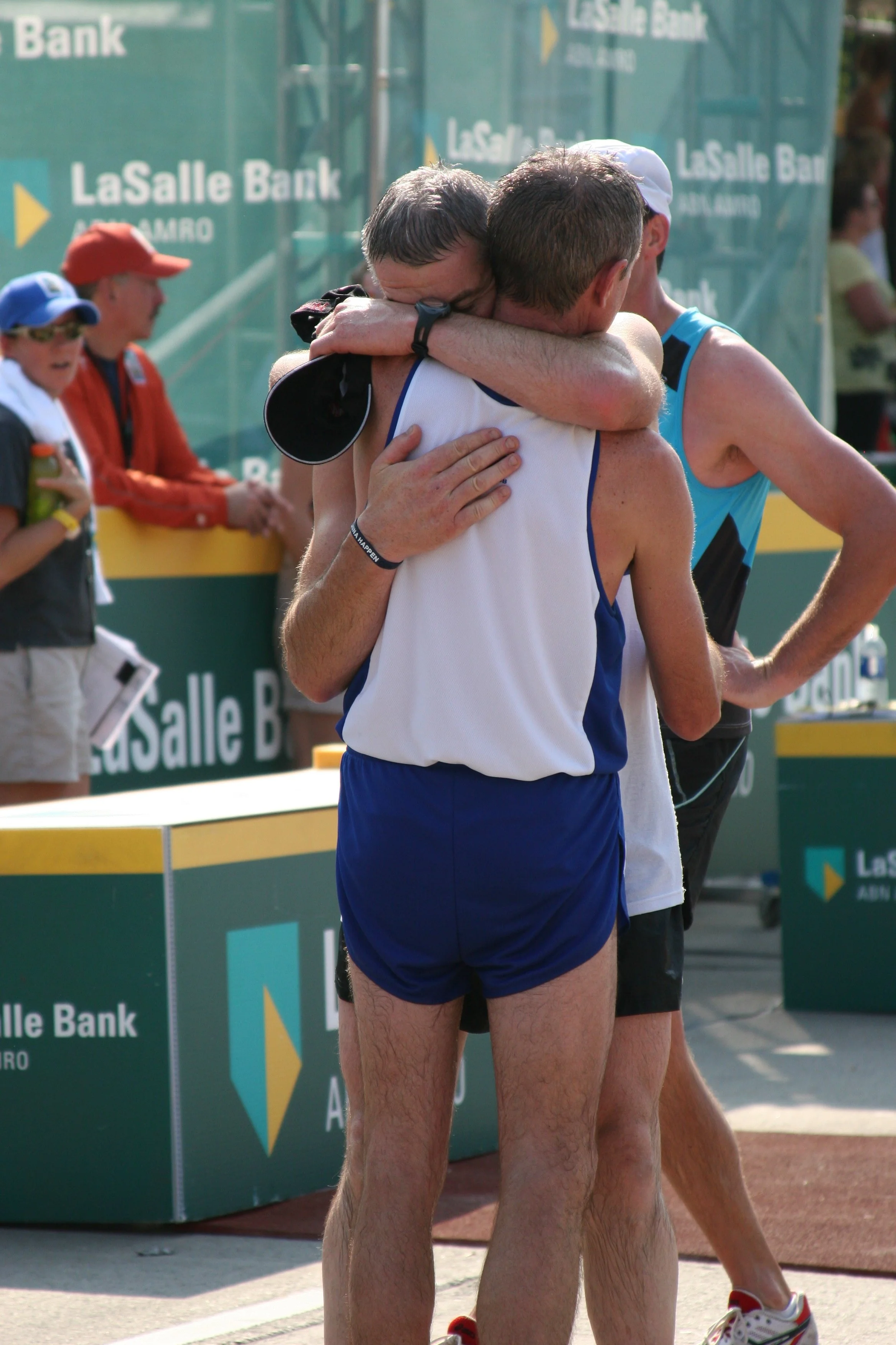Two male runners in athletic gear hugging after finishing a race near a green sponsored backdrop. Spectators watch nearby.