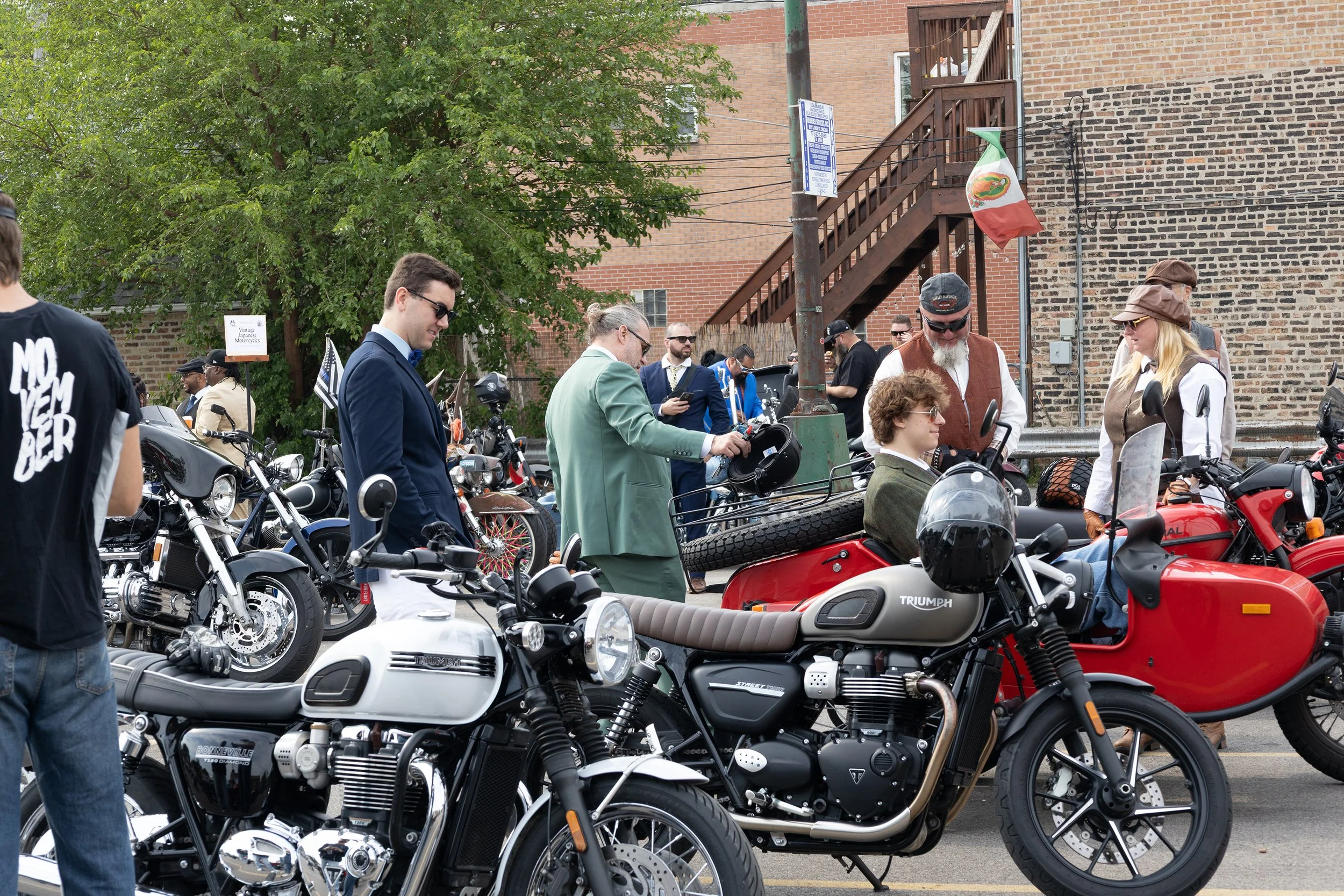 A group of people gathered around vintage and modern motorcycles in an outdoor setting, with some individuals inspecting or conversing. The scene includes a large green tree, brick buildings, and a Mexican flag hanging from a pole.