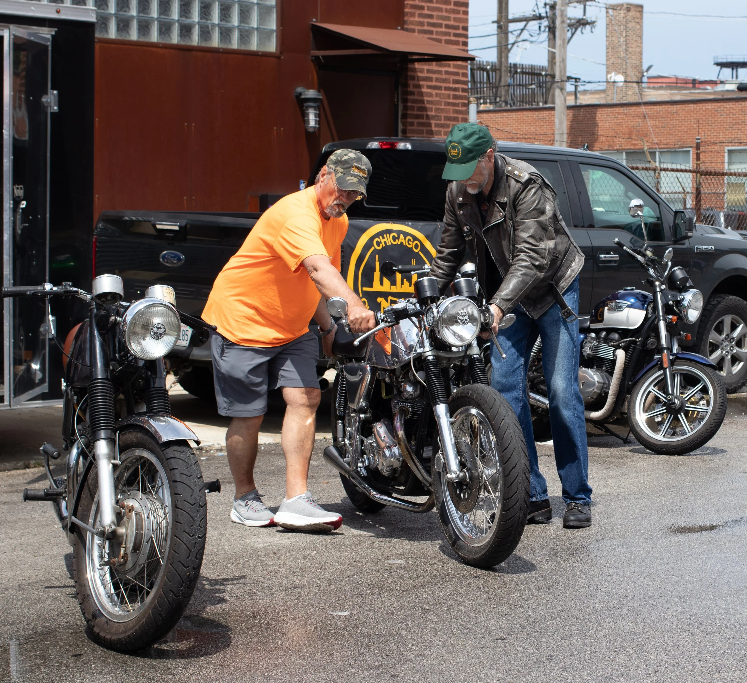 Two older men working on motorcycles outside near a black truck with 