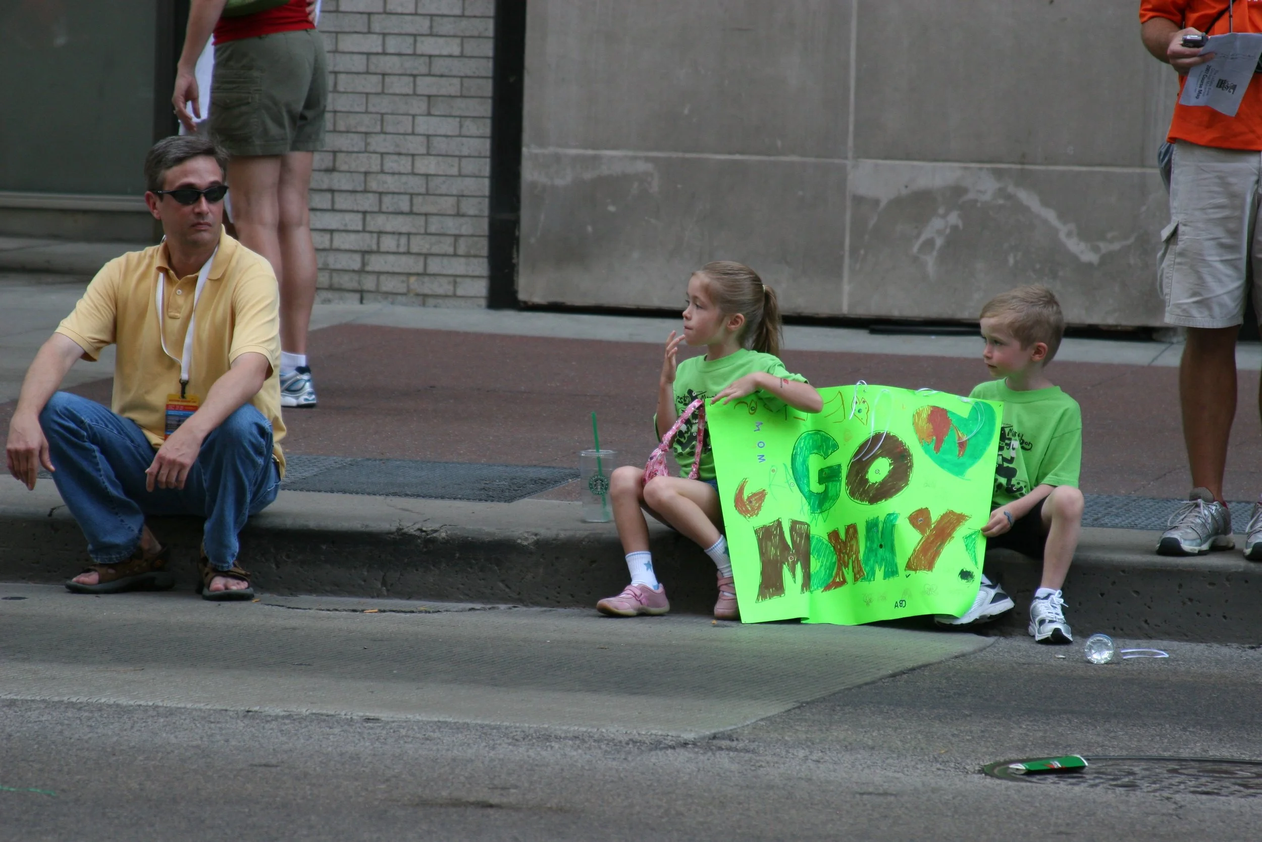 Two children sitting on a sidewalk holding a bright green protest sign that says 'Go Mom!' with colorful decorations. An adult man wearing a yellow polo shirt and sunglasses is sitting nearby. There are other adults standing around, and the setting a