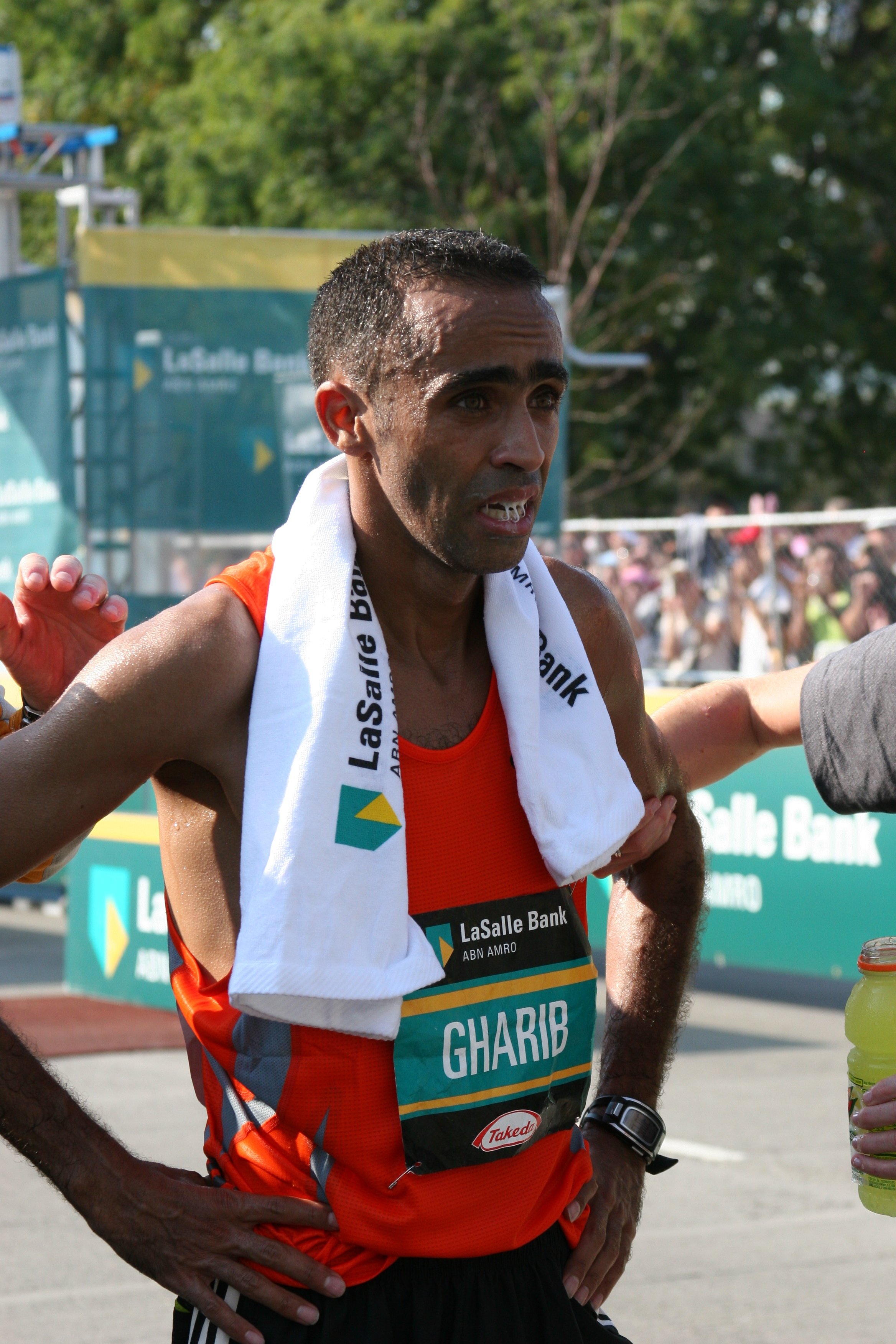 A male marathon runner wearing an orange tank top with a white towel around his neck, standing with hands on hips, during a race with runners and spectators in the background.