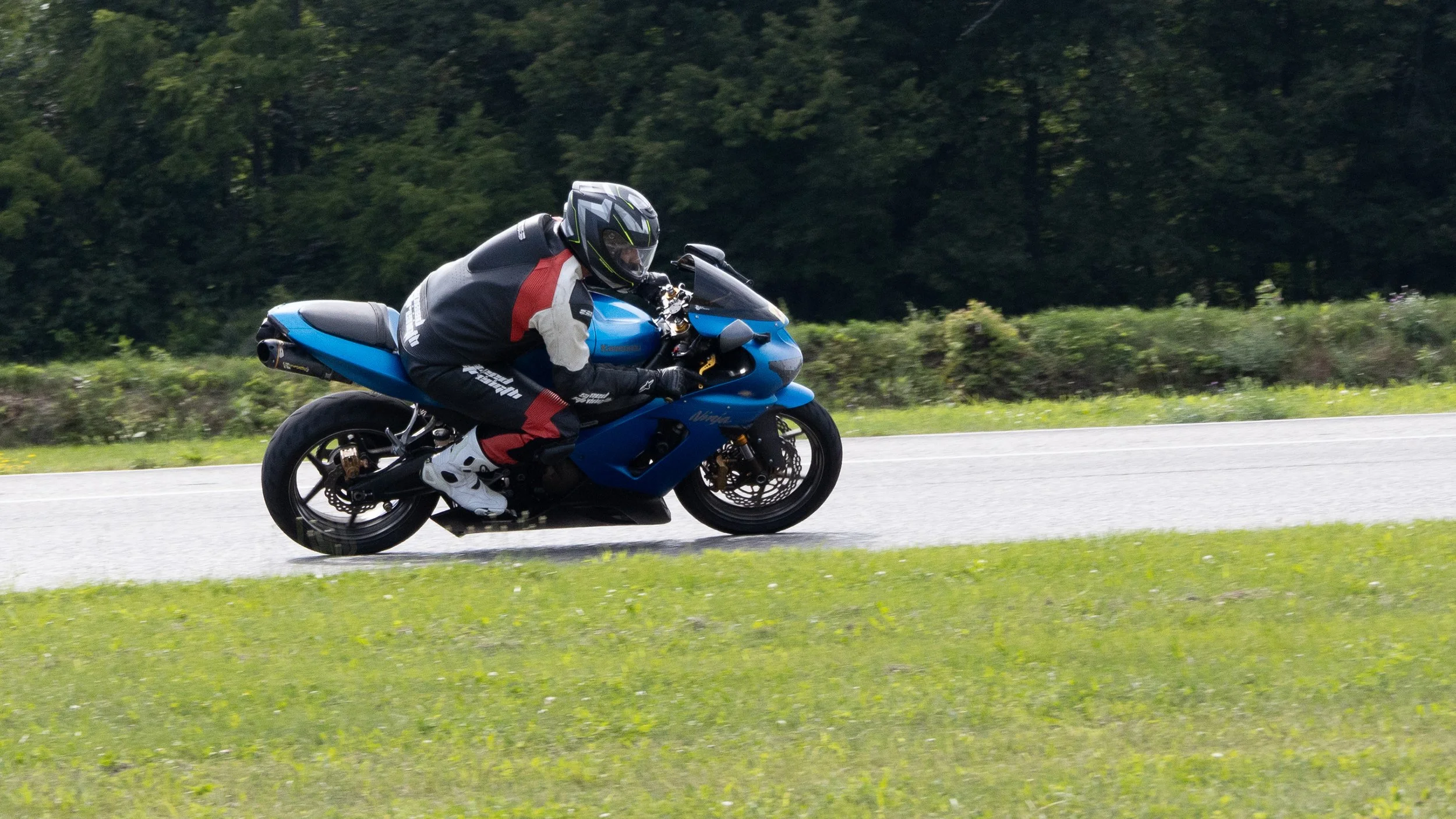 Motorcycle rider in full gear riding on a paved road surrounded by greenery and trees.