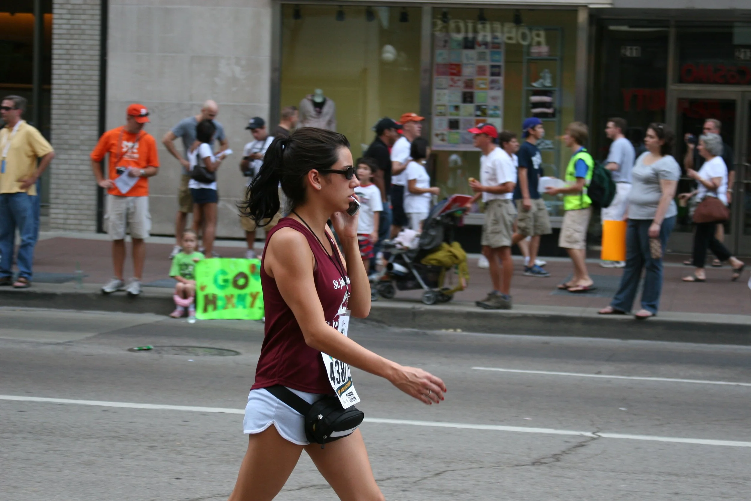 A woman with long dark hair pulled back, wearing sunglasses, a maroon tank top, shorts, and a race bib, is walking across a city street during a public race or marathon. In the background, a group of people, including some children, are gathered outs