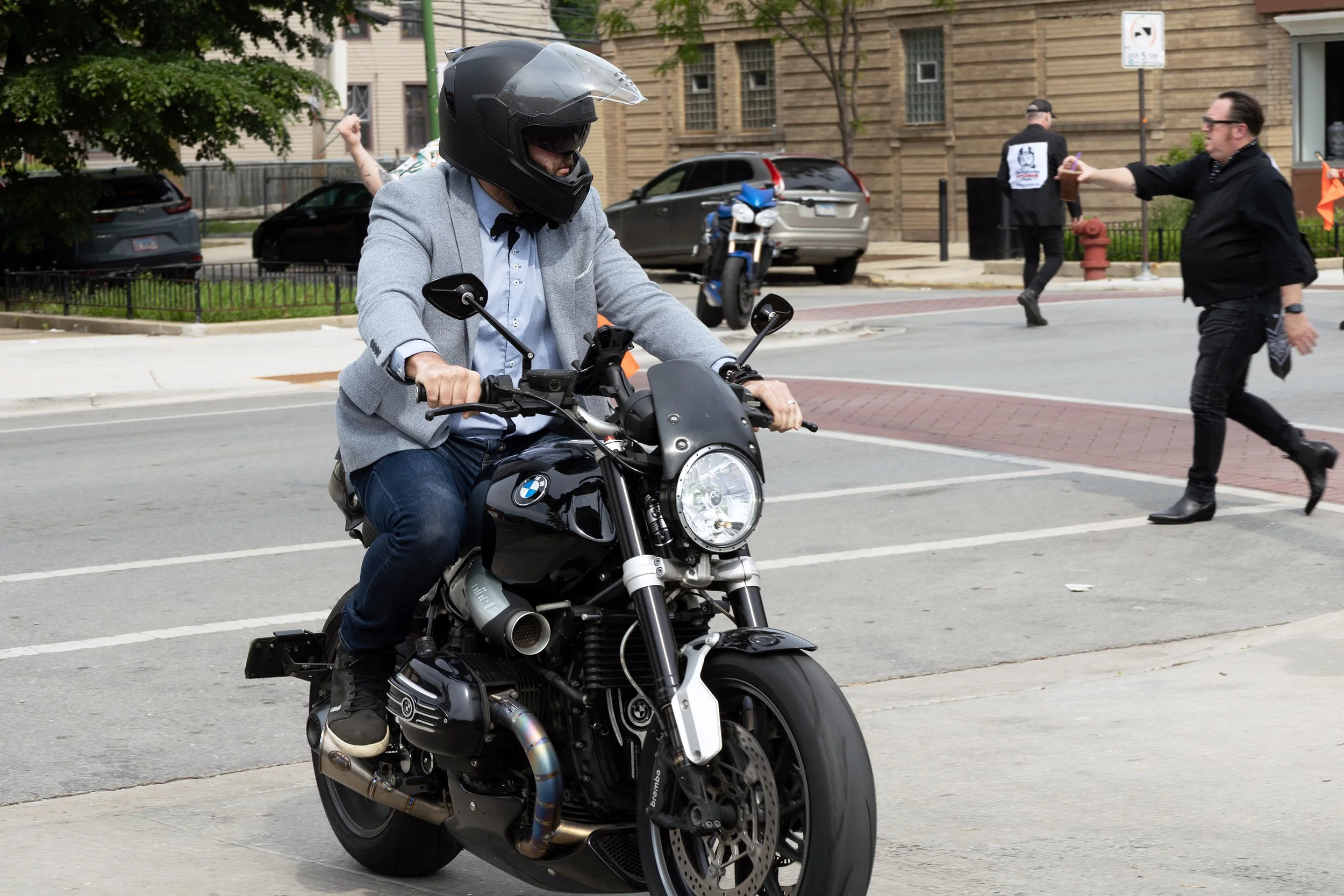 A man wearing a helmet and a grey blazer riding a black BMW motorcycle on a city street.