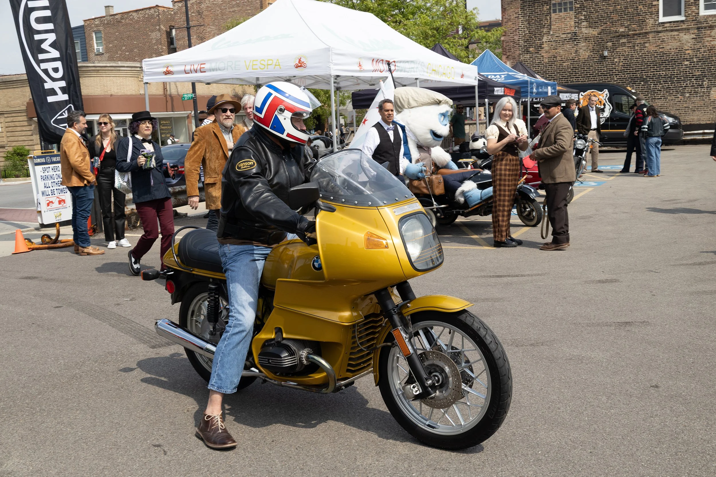 A man in a black leather jacket, jeans, and brown shoes is riding a yellow BMW motorcycle. The horse is carrying a man in a white vest, black bow tie, and checkered pants, and a person in a mascot costume resembling a white horse with blue details is