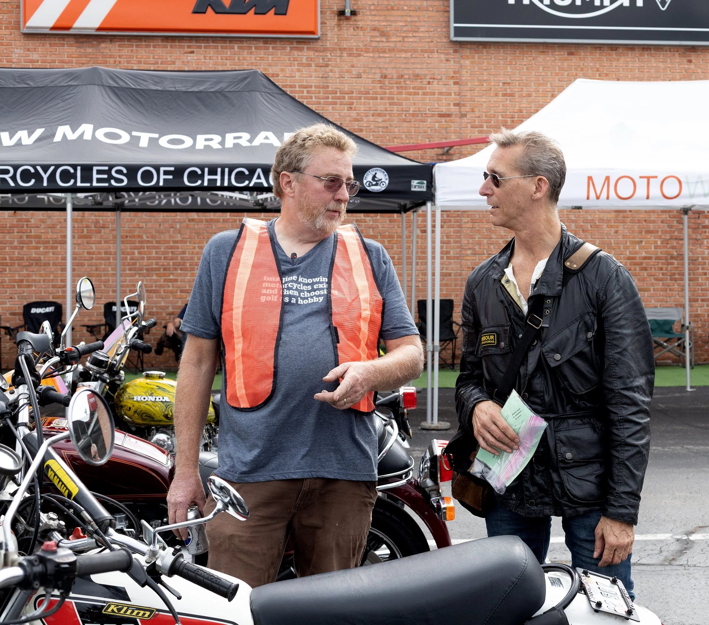Two men are talking at a motorcycle event with motorcycles parked in front of them. One man is wearing a blue t-shirt and orange safety vest, the other is wearing sunglasses and a black leather jacket. There are tents and a brick building in the back