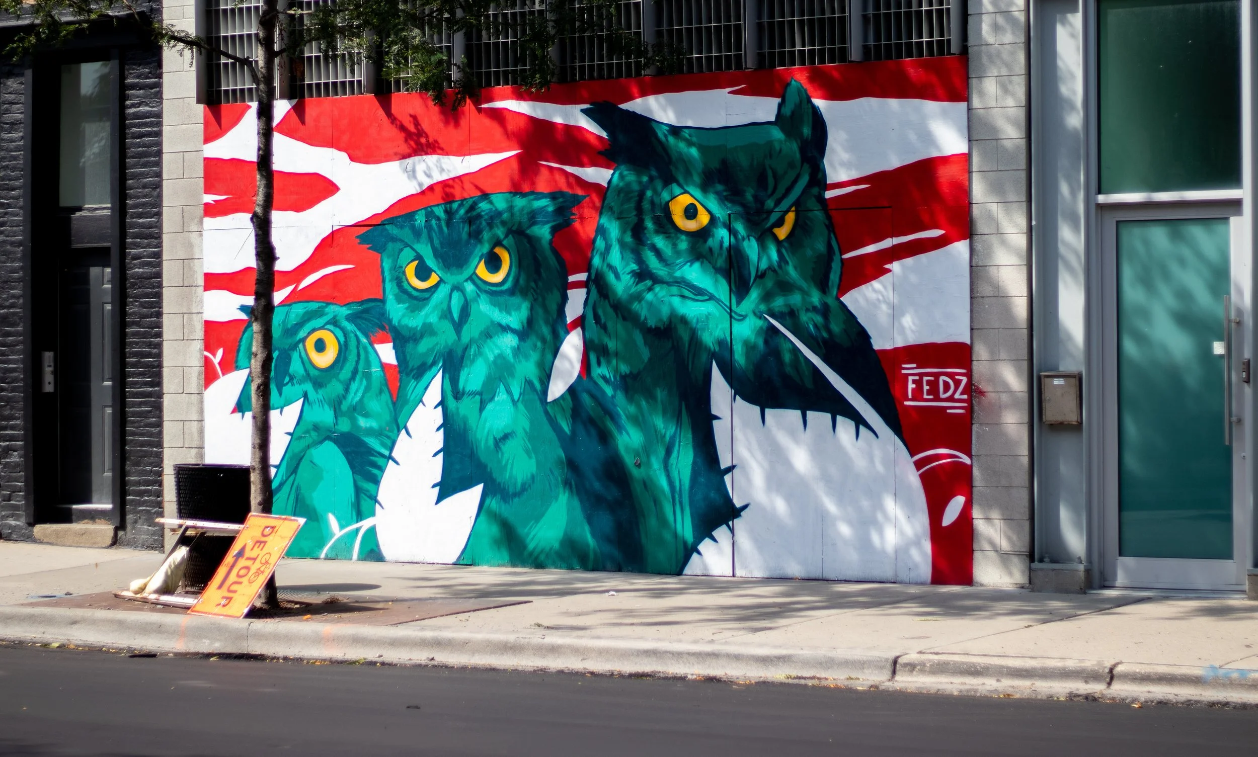 Street art mural of three stylized owls in shades of blue and green on a building side, with red and white abstract background, in an urban setting.