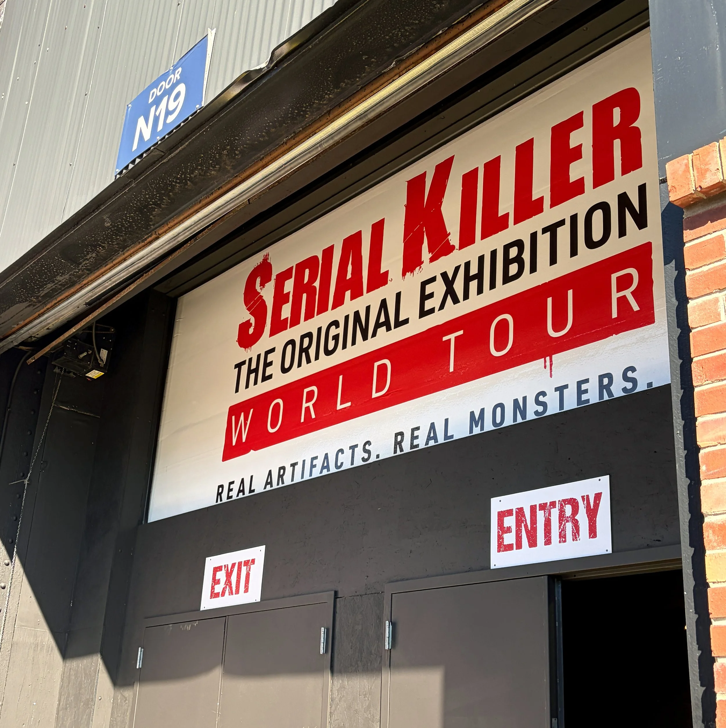 The image shows a sign on a building indicating an ongoing sale of artifacts, with smaller signs for fire exit doors underneath. The building has a brick column on the side and the signs are mounted under a black overhang.