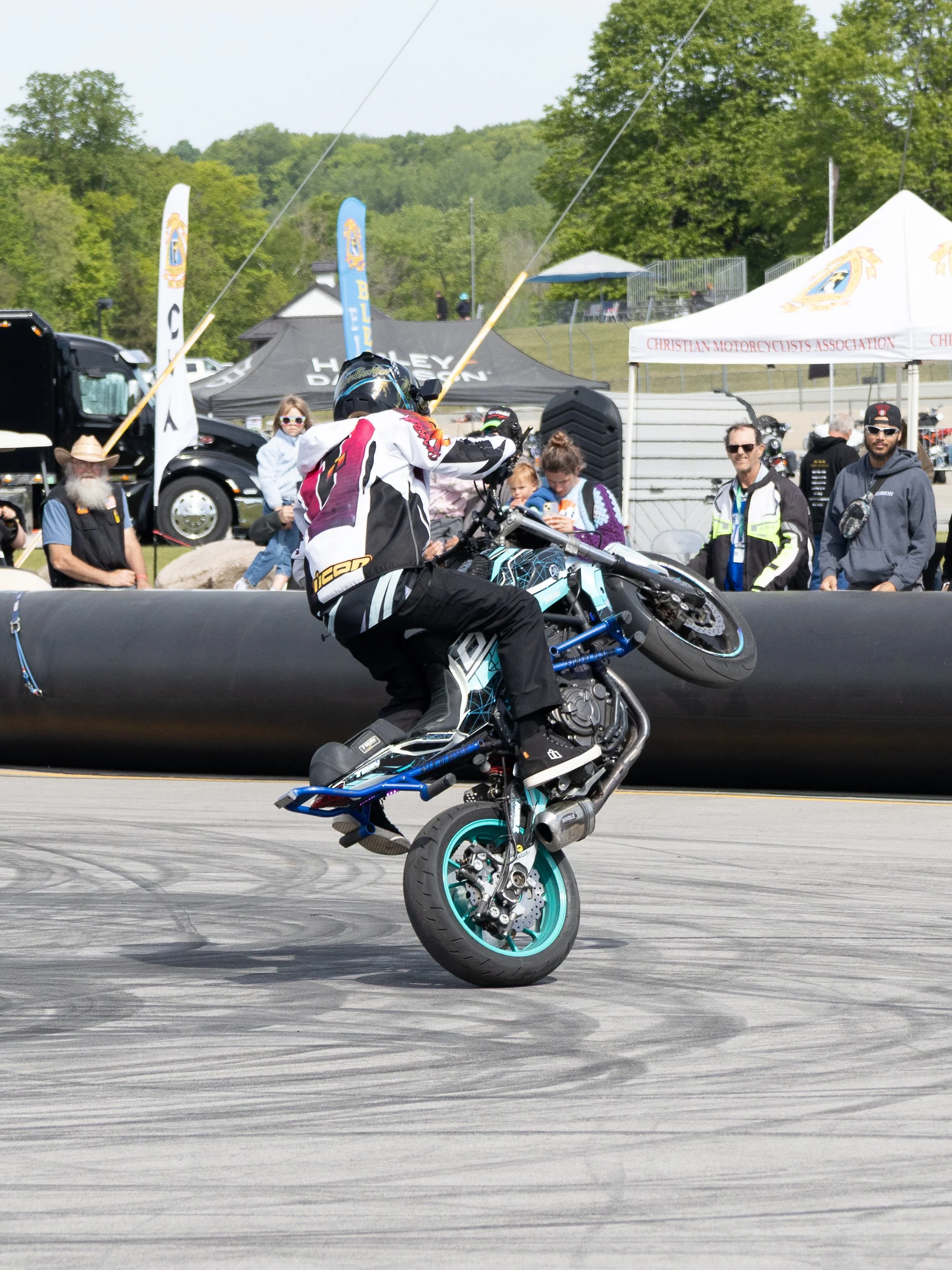 A motorcyclist performing a wheelie stunt on a blue and black motorcycle at an outdoor event, with spectators and tents in the background.